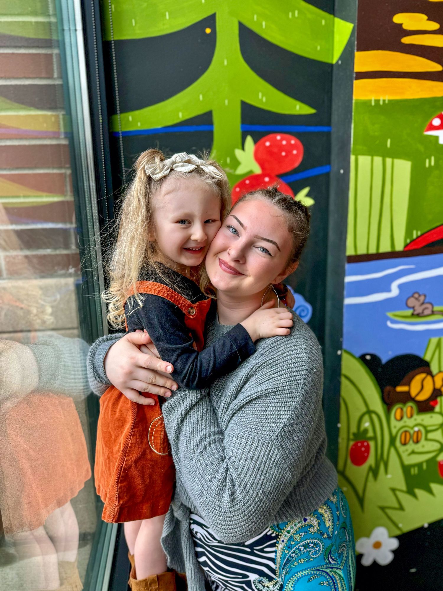 A young woman smiles warmly while holding a toddler girl in an orange dress in front of a colorful mural.