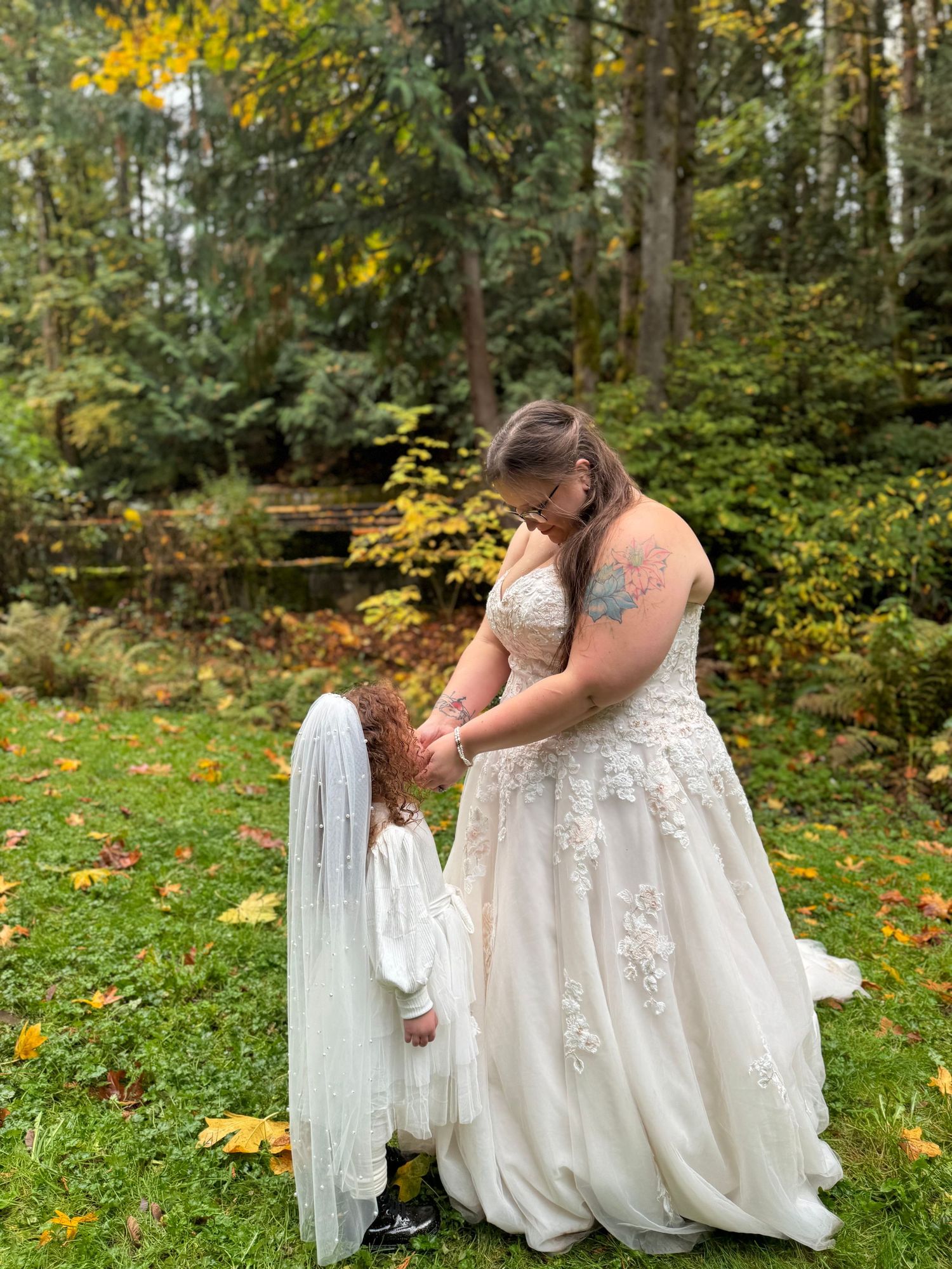 Bride in lace gown adjusting flower girl veil in autumn forest setting.