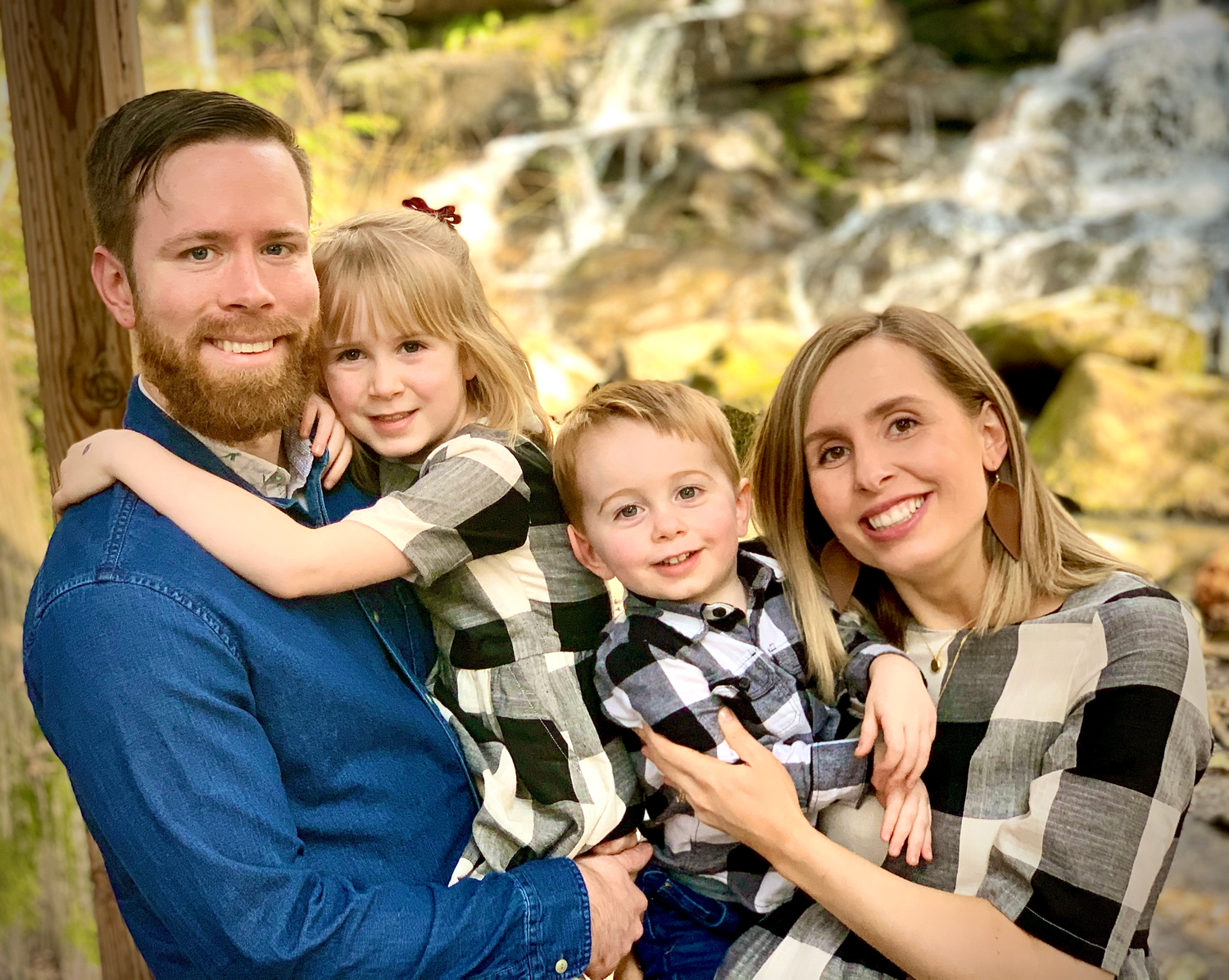 Joyful family of four posing in front of a waterfall, parents holding two young children in matching outfits.