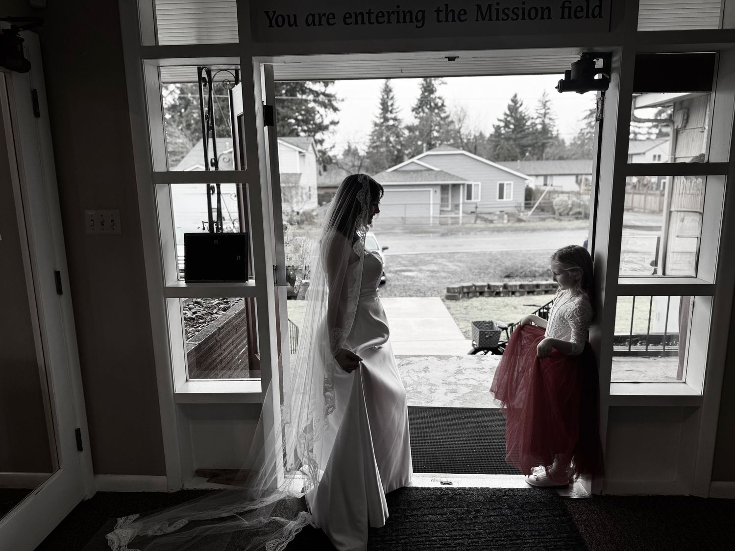 Black and white photo of a bride in a white gown standing in a doorway with a young girl in a red dress.