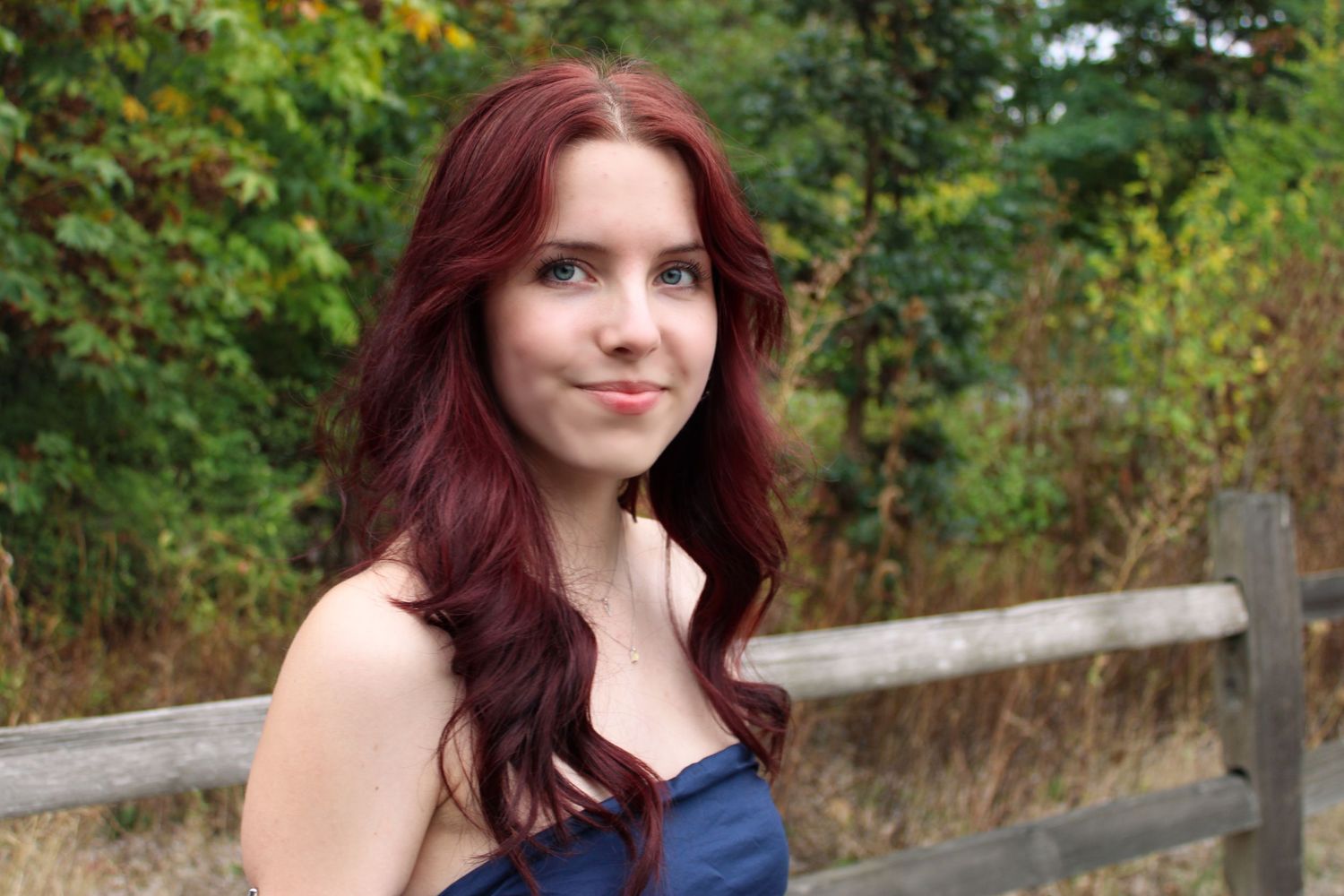 A young woman with long red hair smiles outdoors near a wooden fence in fall.