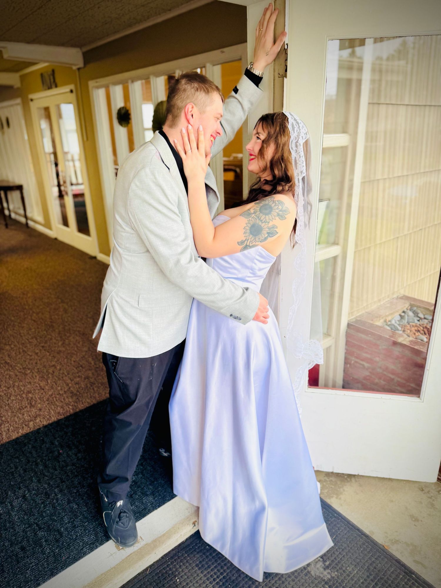 Bride in a lavender gown with tattoos and veil shares a romantic moment with groom in a beige blazer.