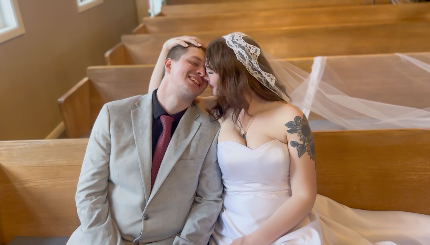 A bride and groom sit closely together on a wooden church pew sharing a tender romantic moment.