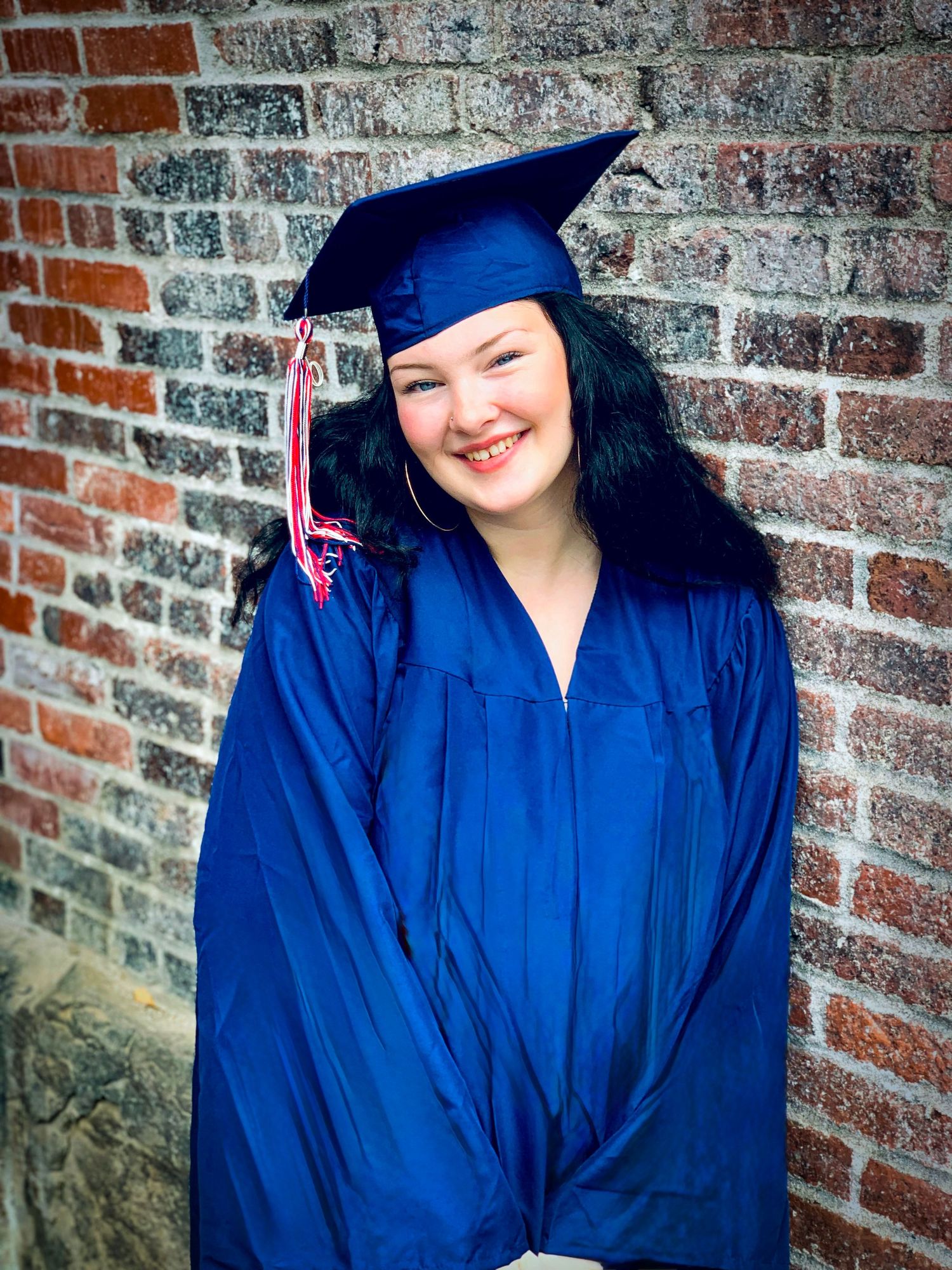 Smiling young woman in blue graduation cap and gown posing against a brick wall at graduation ceremony.
