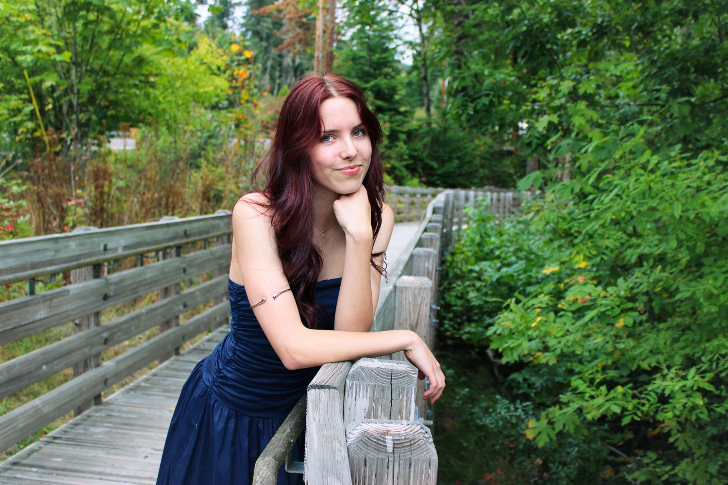 Young woman in navy strapless dress leans on wooden bridge railing surrounded by green foliage.