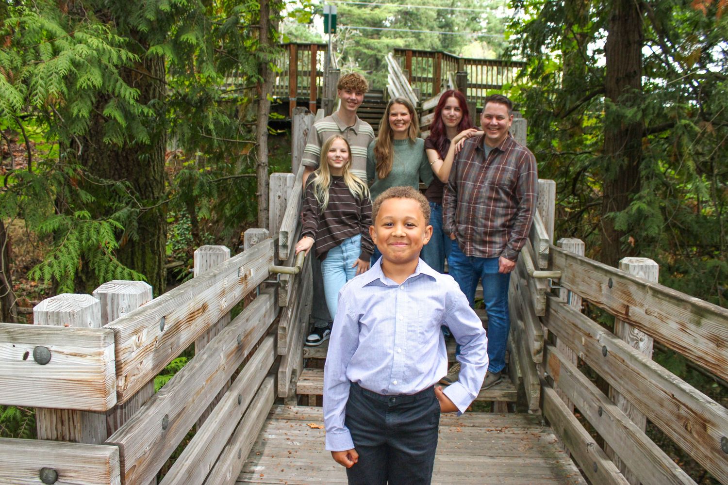 A young boy in a blue shirt stands confidently on a wooden bridge with his family posed behind him in a lush forest.