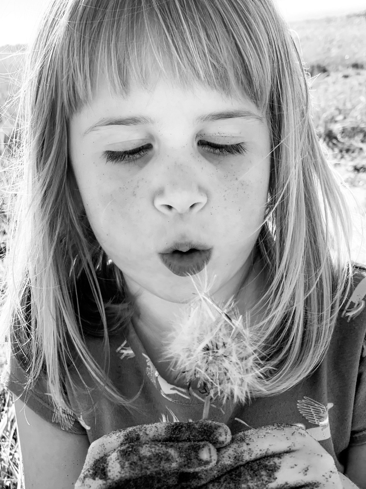 Black and white photo of young girl with freckles blowing a dandelion flower.
