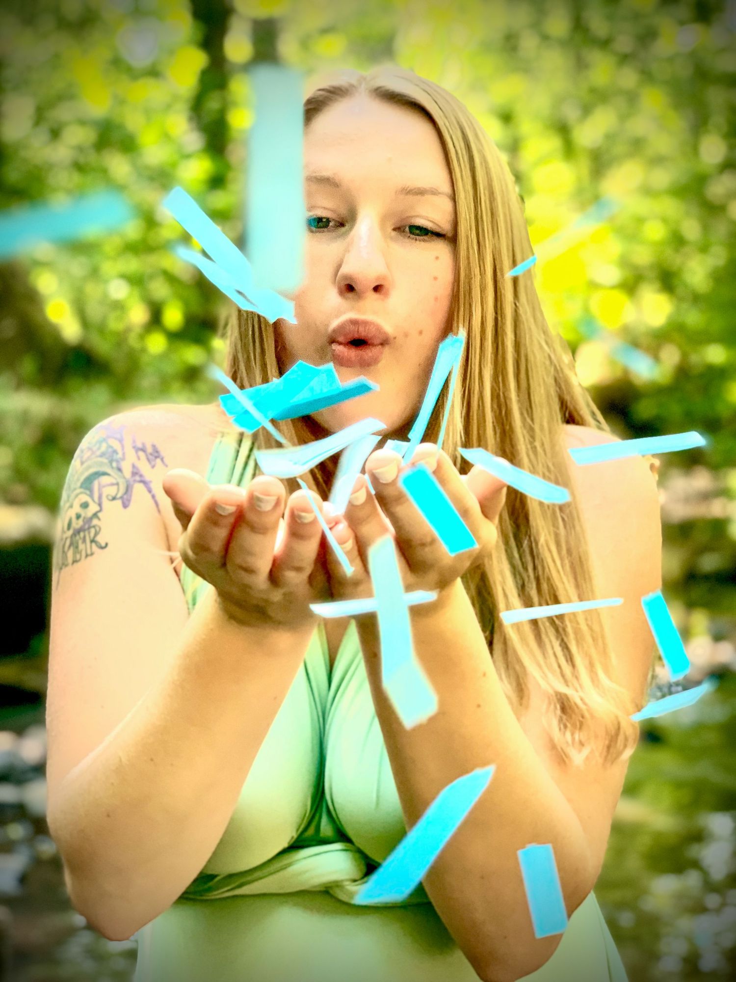 Young blonde woman blowing blue confetti from her hands outdoors in a garden.