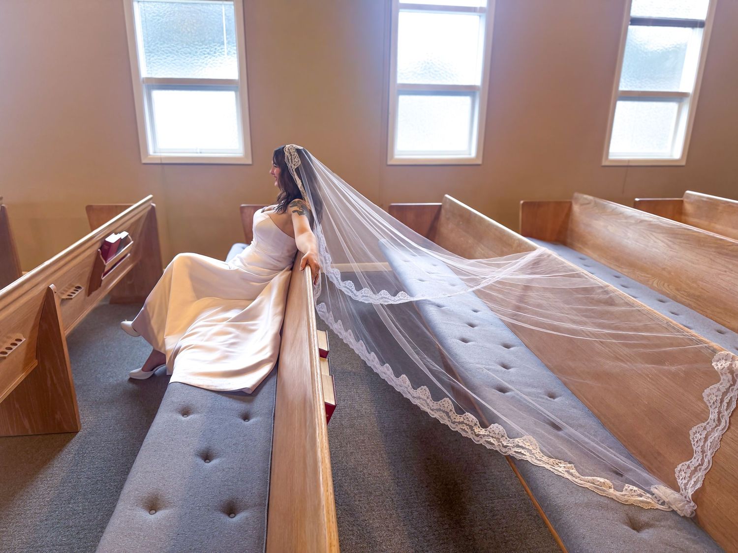 Bride in a white wedding dress with an elegant long lace-trimmed cathedral veil draped across church pews.