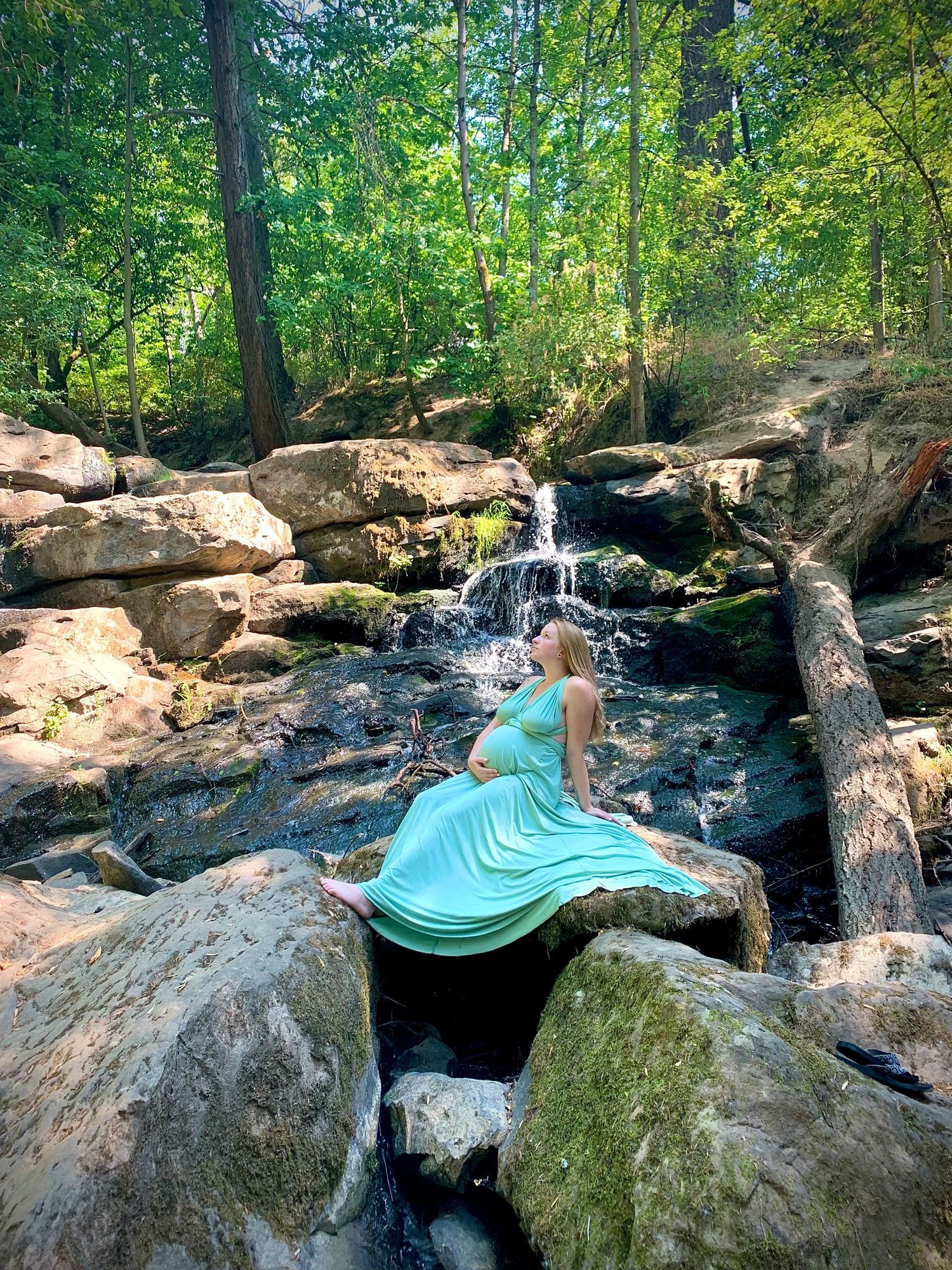 Pregnant woman in a flowing mint green dress sitting on rocks near a small waterfall in a lush forest.
