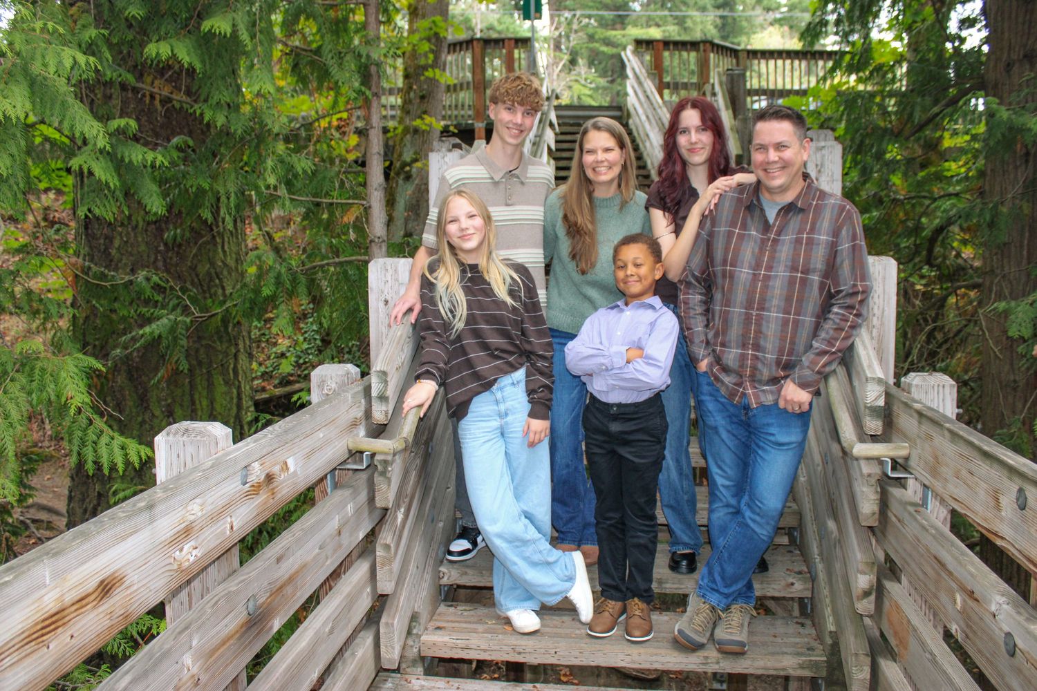 Family of six poses together on wooden outdoor staircase surrounded by green trees.