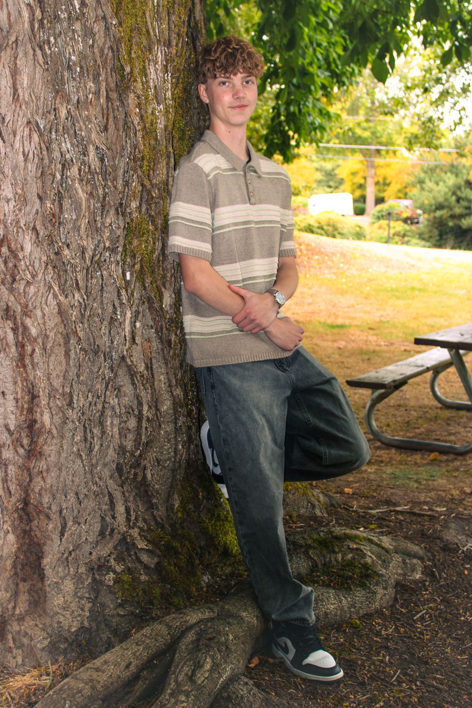 A teenage boy in a striped polo shirt leans casually against a large moss-covered tree in a park setting.
