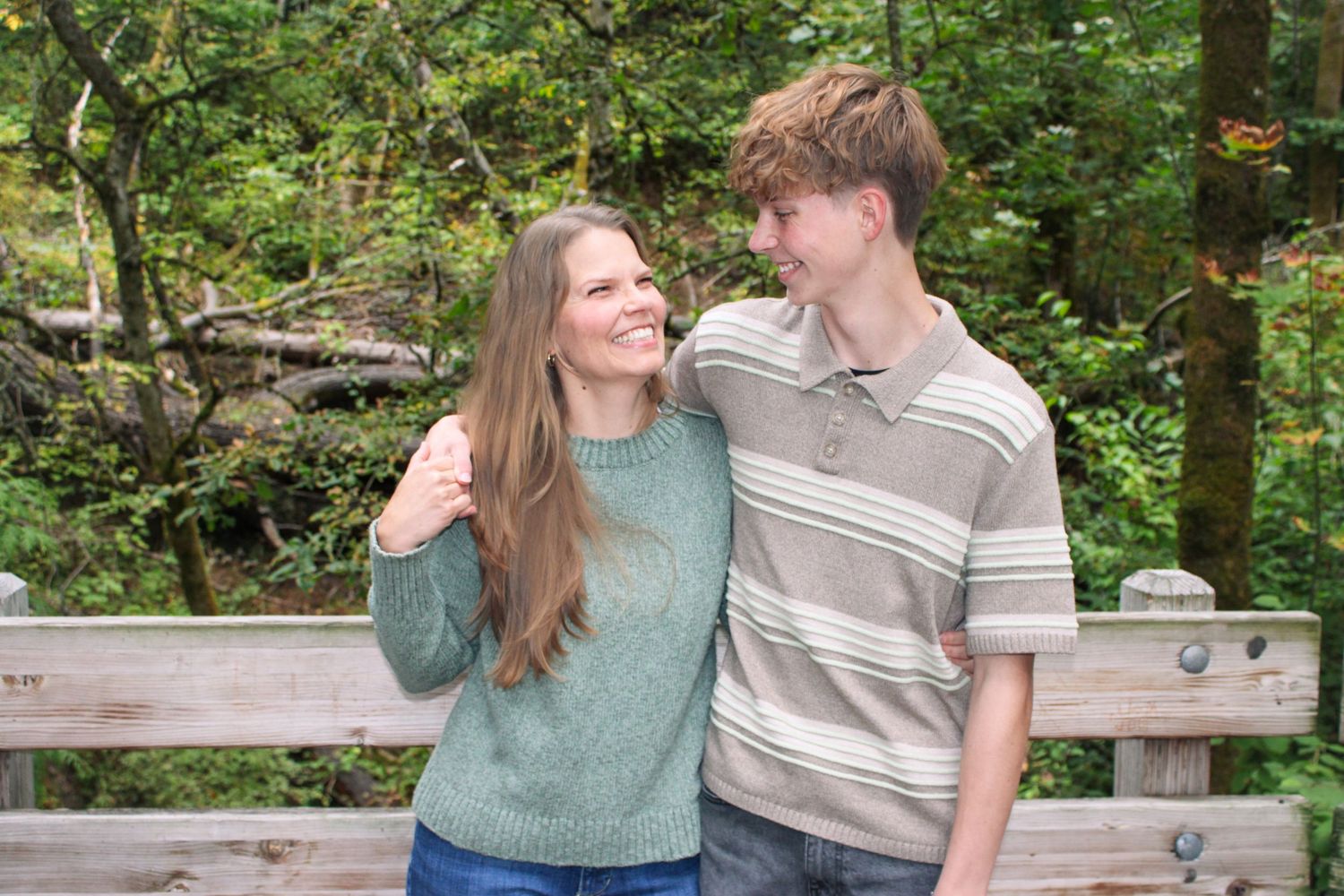 Smiling woman in green sweater and tall teen boy pose together on wooden bridge outdoors.