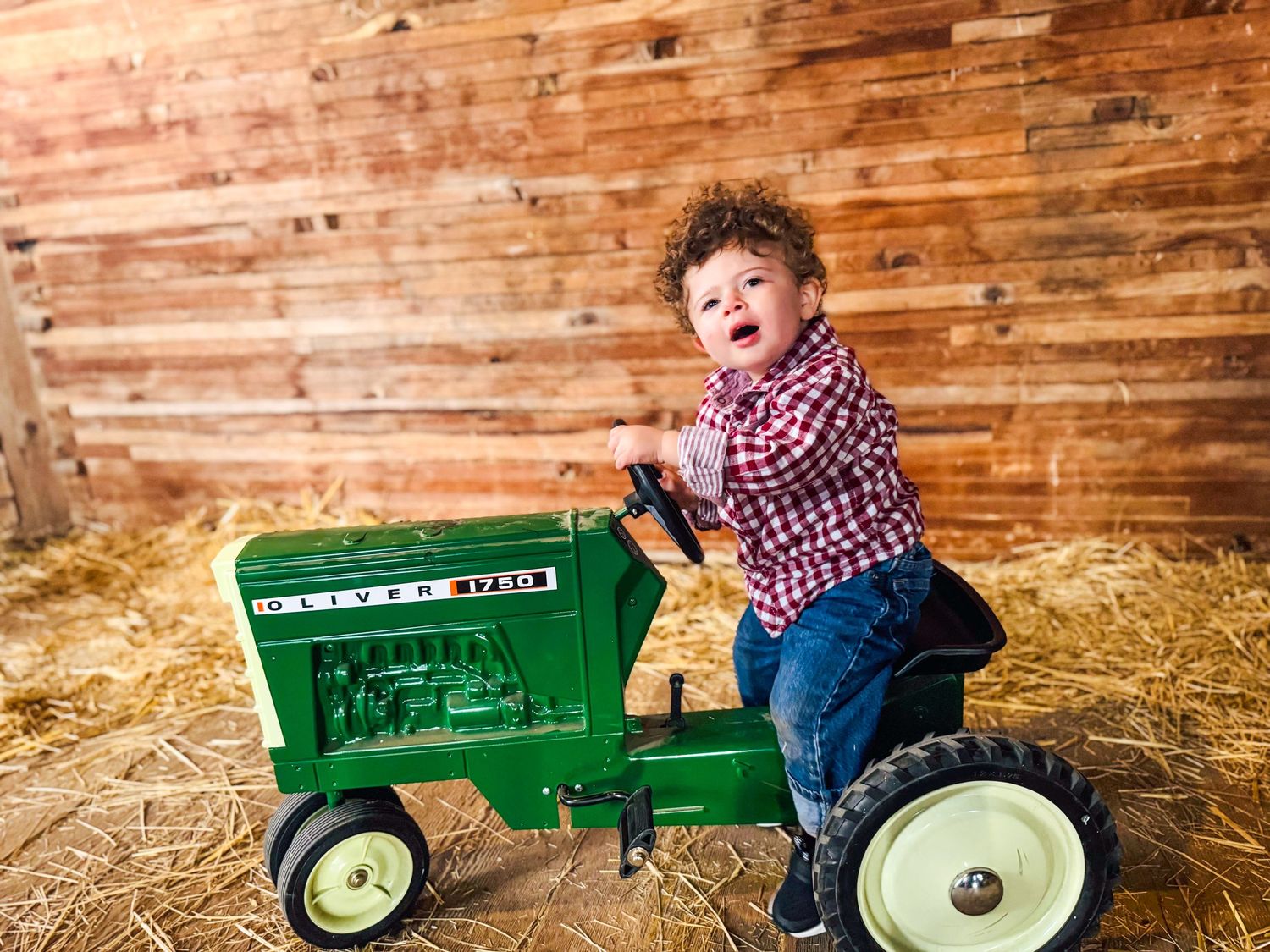 A curly-haired toddler in a plaid shirt sits on a green Oliver pedal tractor in a rustic hay-filled barn.