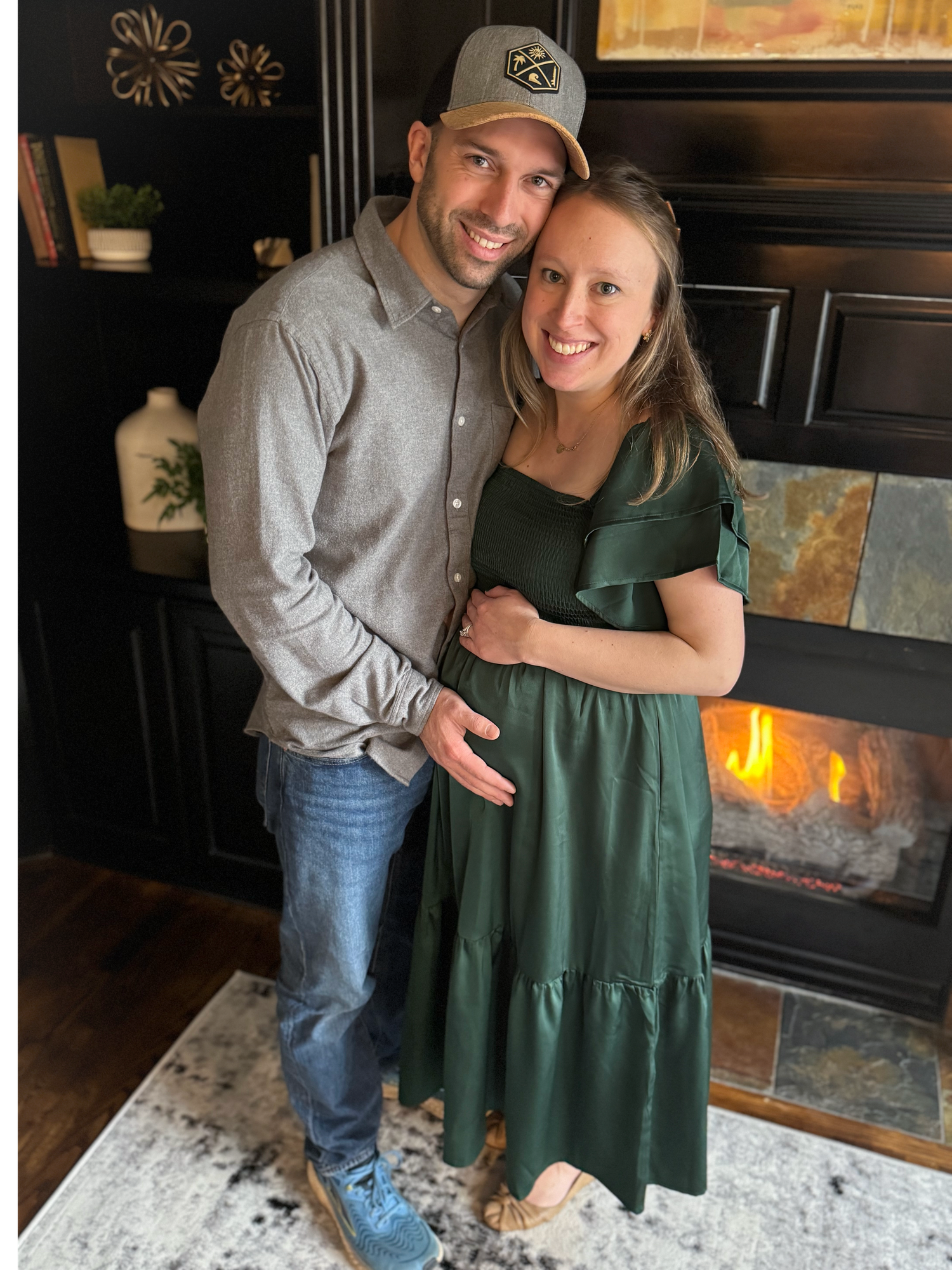 Smiling couple posing together in front of a fireplace, woman in green maternity dress, man in gray shirt.