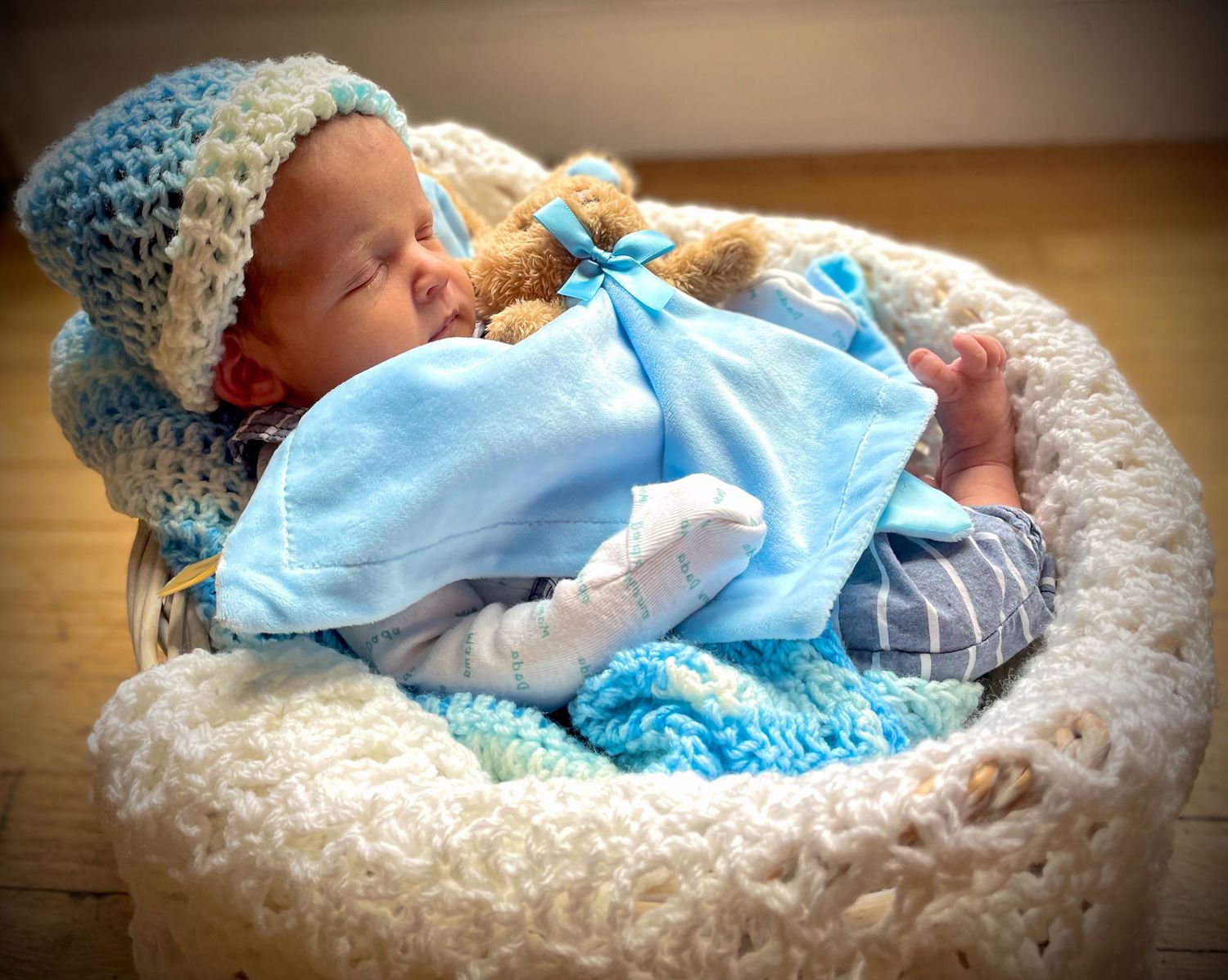 A sleeping newborn baby dressed in blue rests peacefully in a crocheted basket with a small teddy bear.