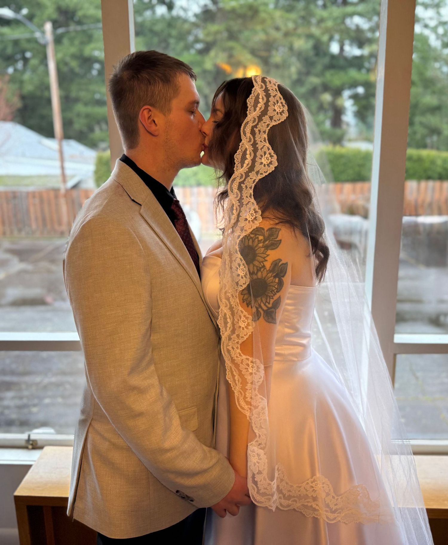 Newlywed couple shares a tender kiss by a large window, bride wearing a lace veil and groom in a beige suit.