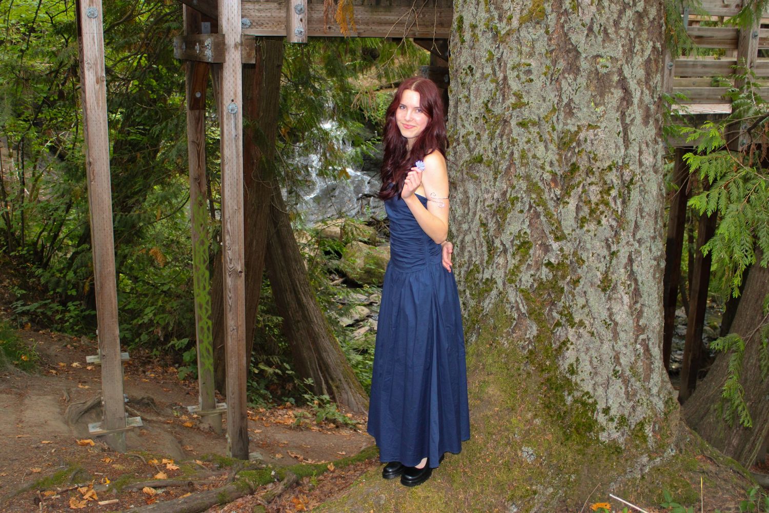 Young woman in navy blue formal gown stands beside large tree in wooded outdoor setting.