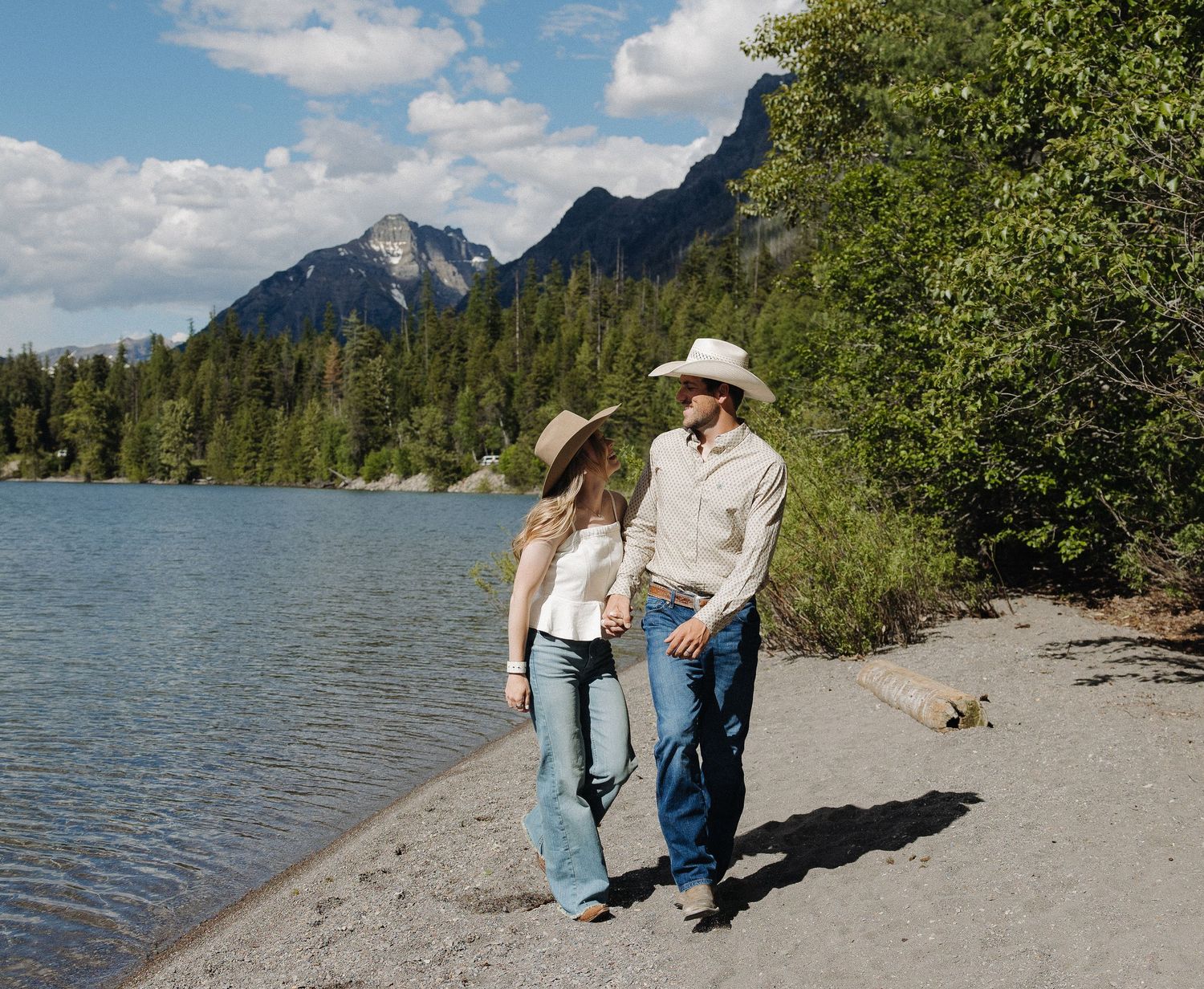 Couple in cowboy hats and jeans standing by a mountain lake shore with pine trees and peaks.