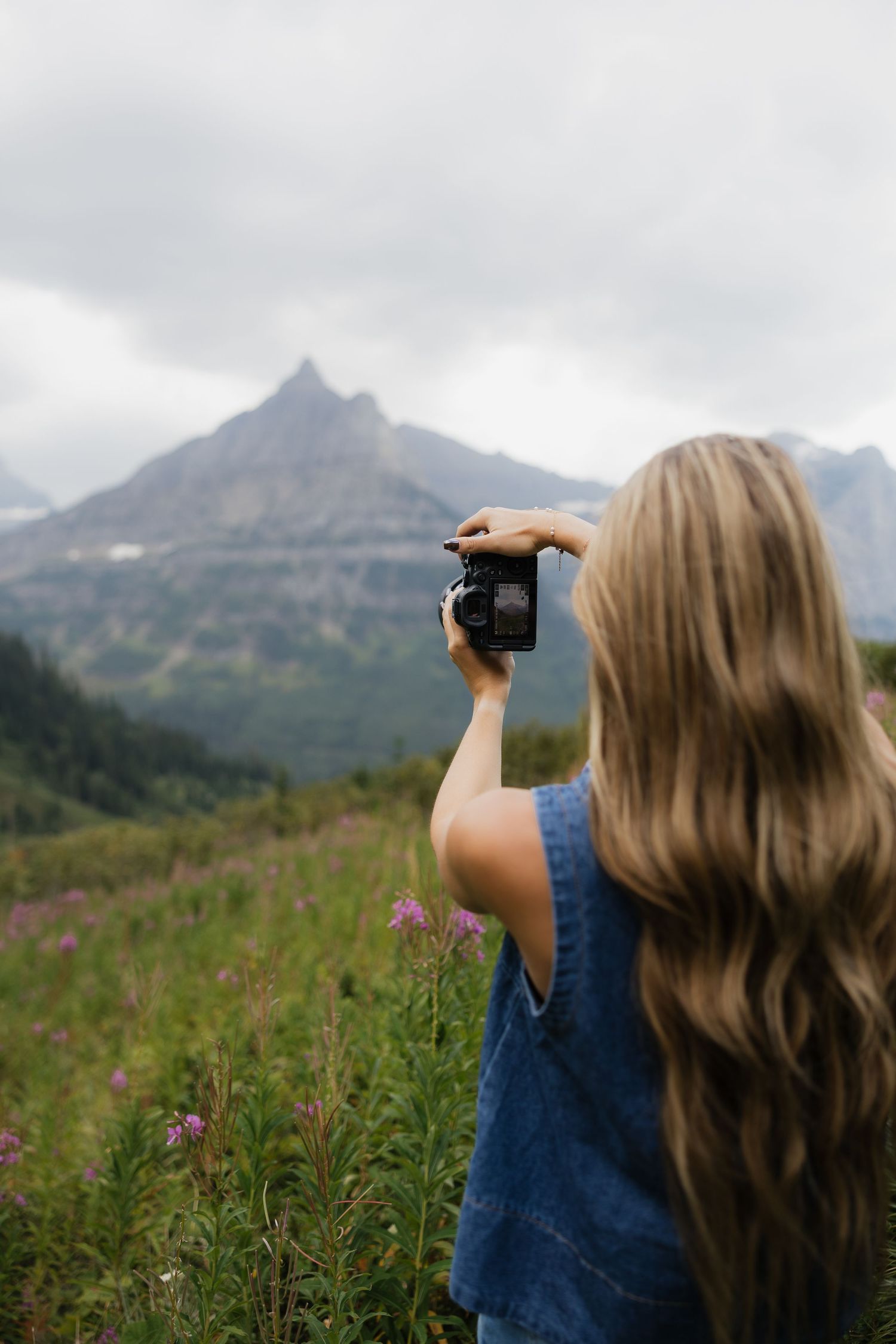 Blonde woman photographing mountain landscape with wildflowers on cloudy day.