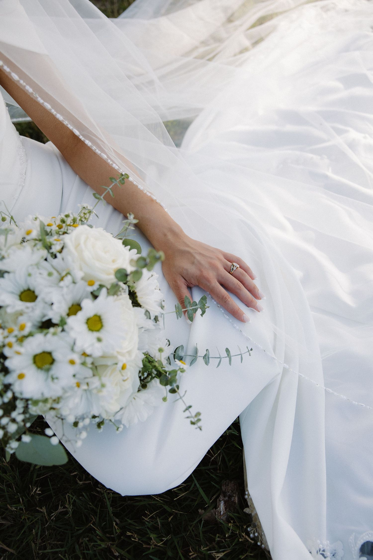 Bride in white gown with veil holding bouquet of white daisies and roses, showing engagement ring on hand.