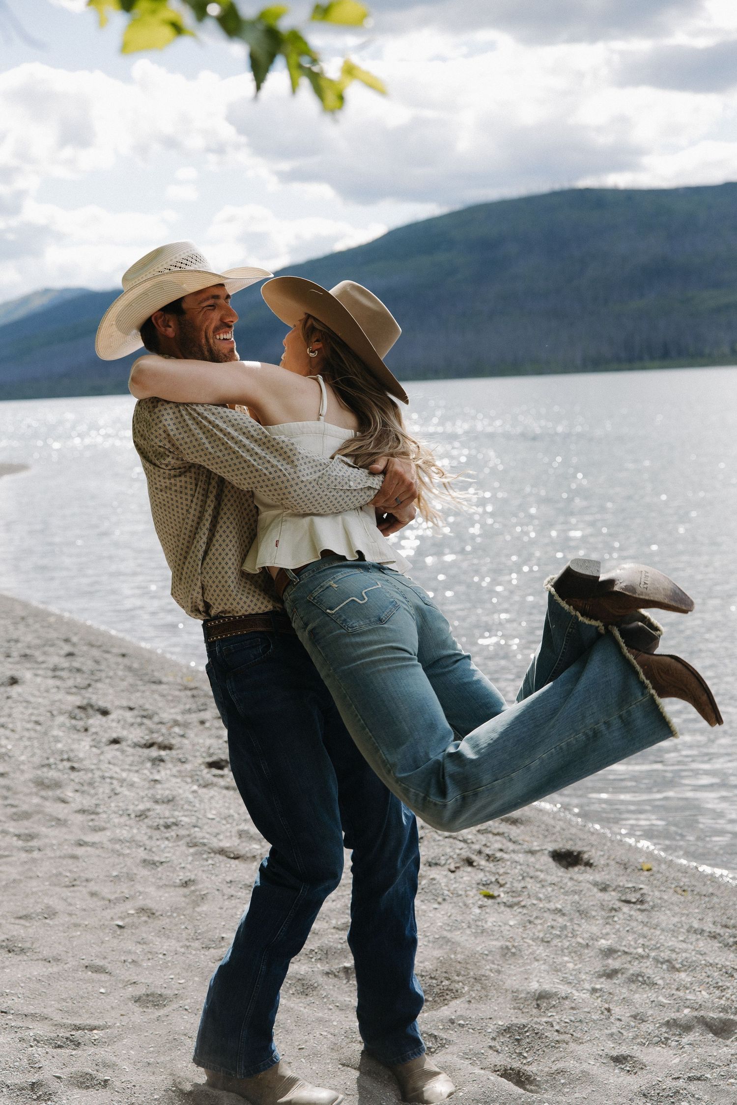 Man in cowboy hat lifting woman on sandy lakeside beach with mountains and water behind them.
