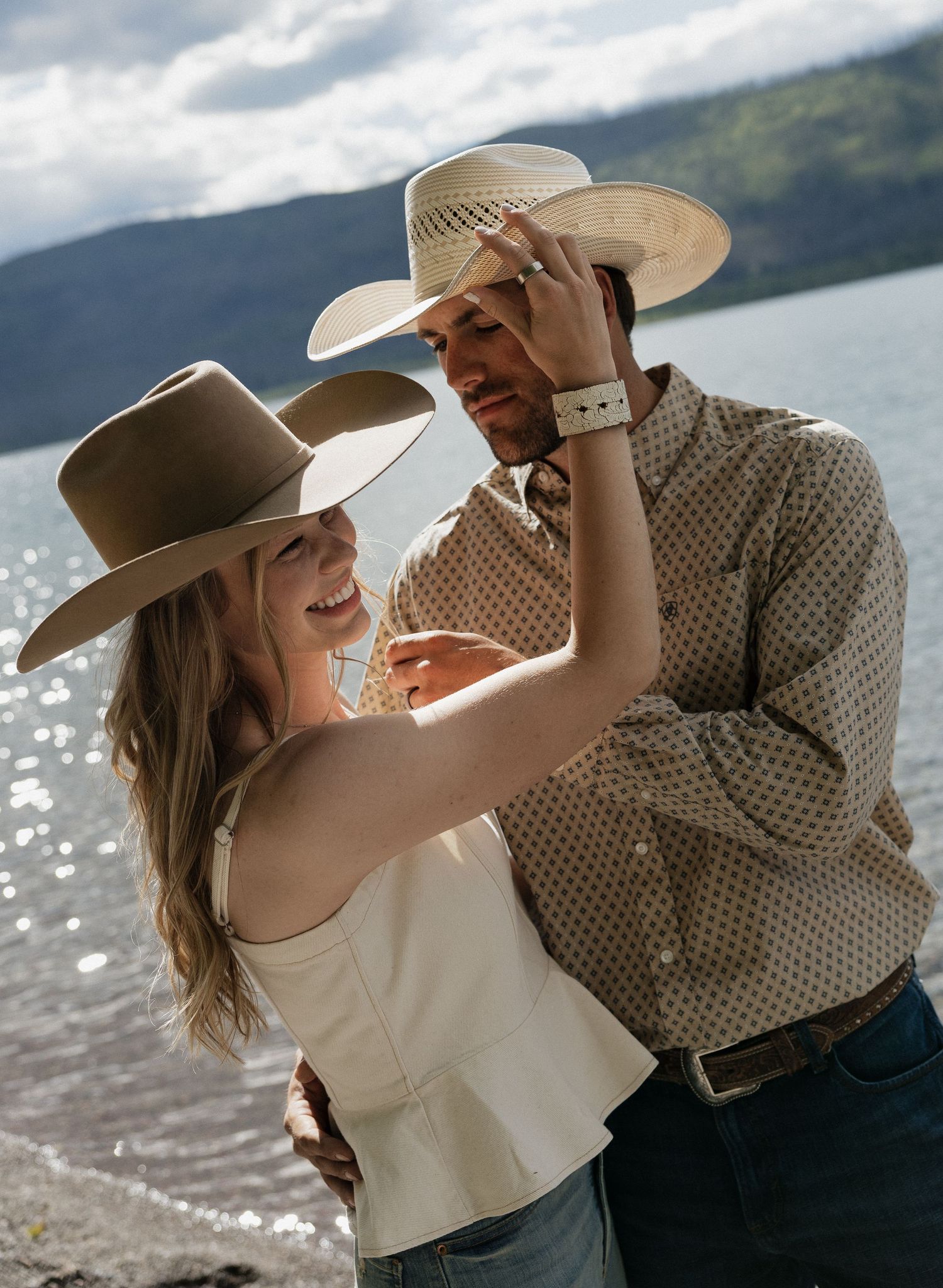 Couple in cowboy hats laugh and dance together near scenic mountain lake shoreline.
Great Falls Montana Couples photoshoot, photographer