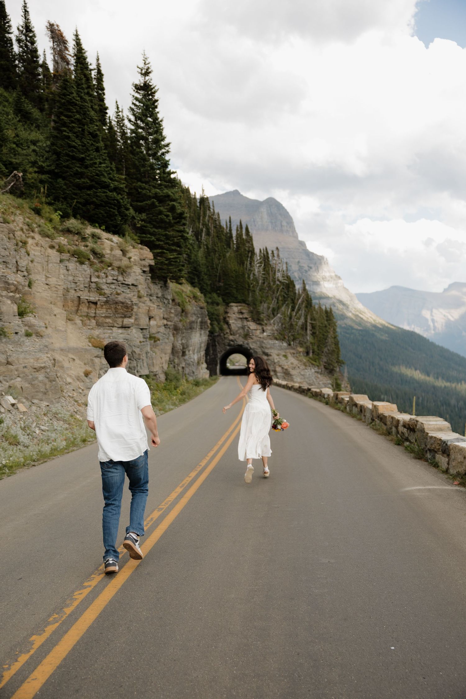 Couple walking on scenic mountain road toward tunnel, surrounded by pine trees and rocky peaks.