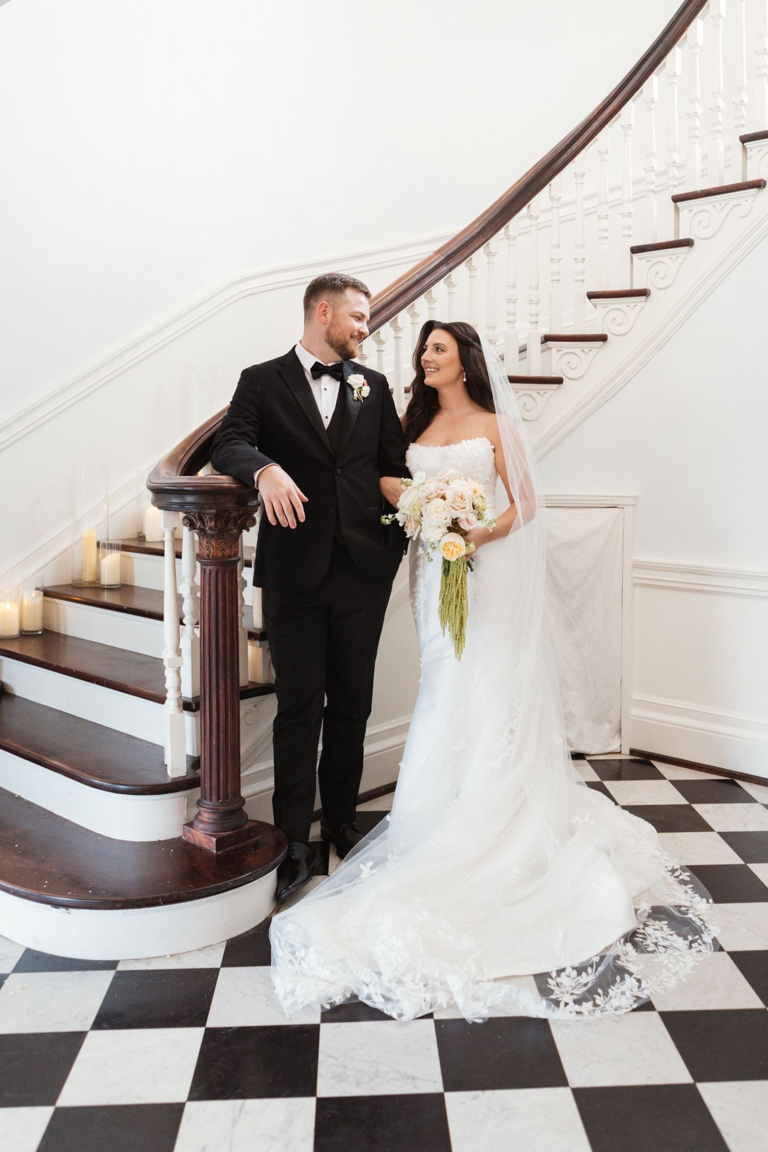 Bride and groom pose elegantly by a curved staircase on a black and white checkered floor in a grand venue.