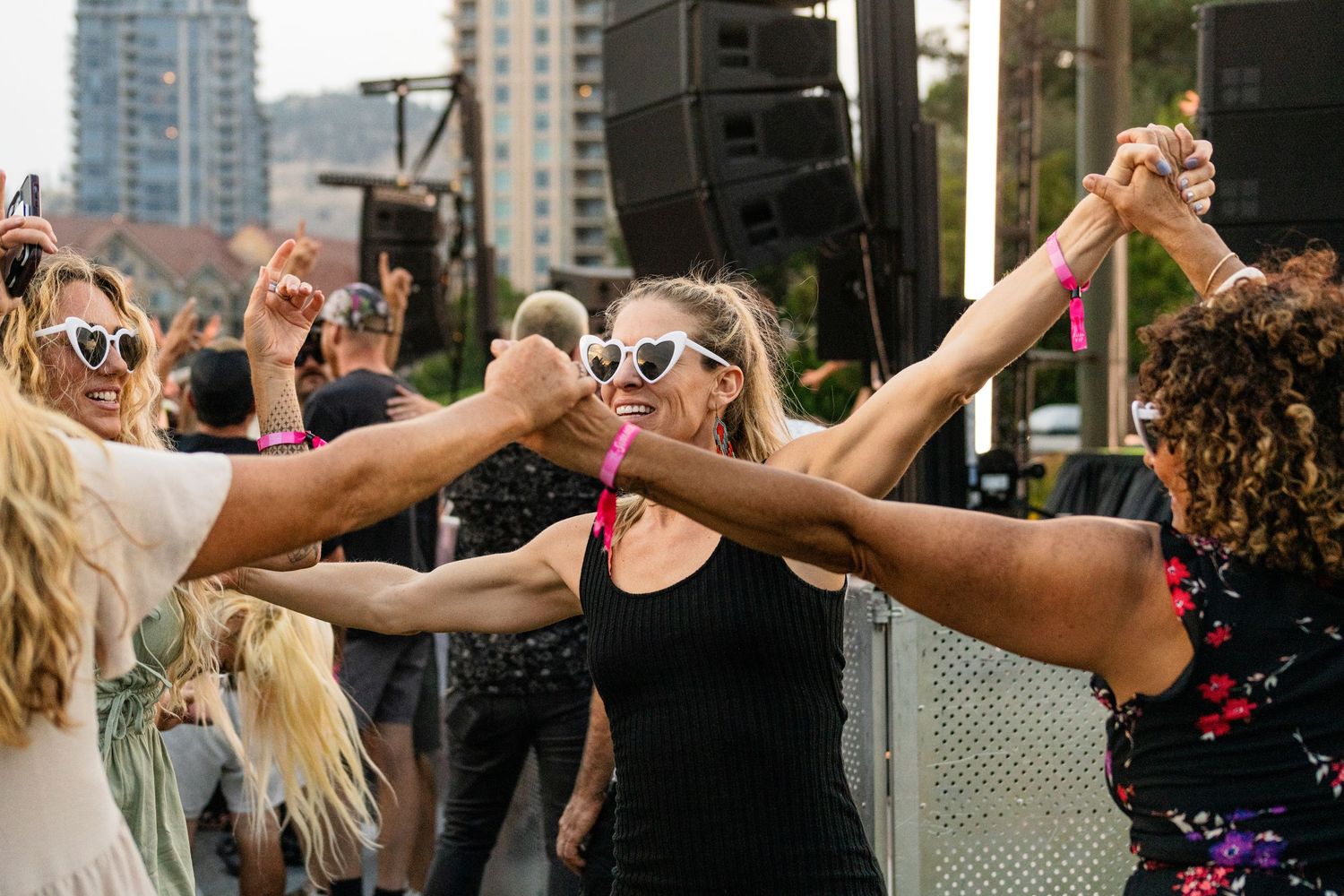 Women dancing and holding hands at an outdoor music festival with speakers and city skyline in background.