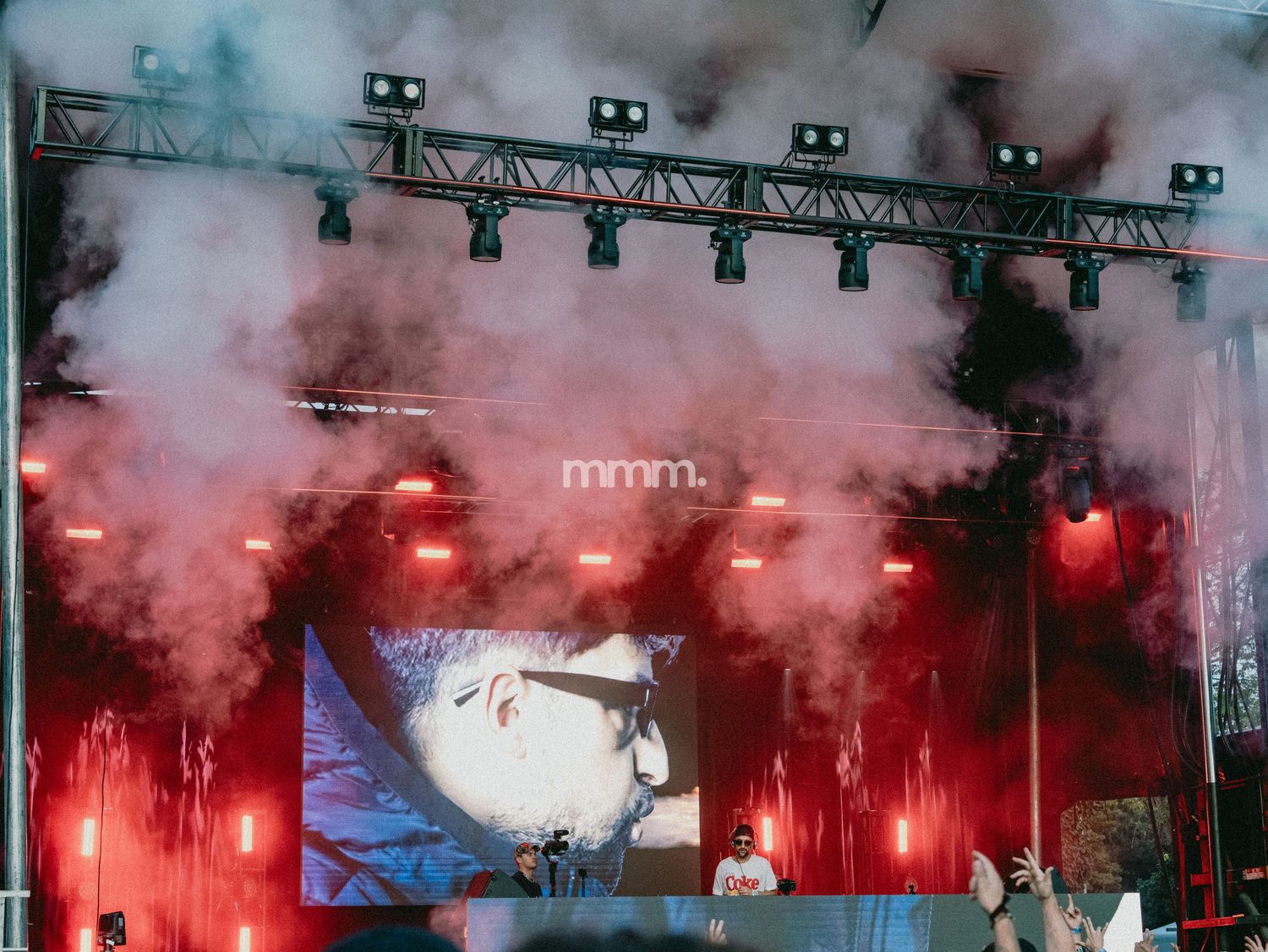 Outdoor festival stage with red lights, smoke effects, and DJs performing under a truss lighting rig at night.