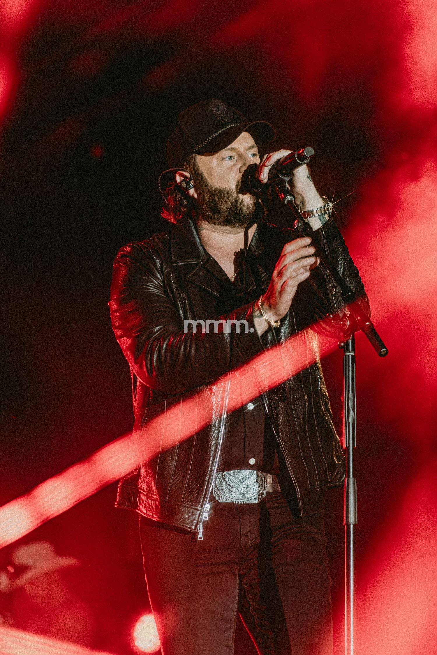 A bearded male singer performs on stage under dramatic red lighting, wearing a black leather jacket and cap while holding a microphone.