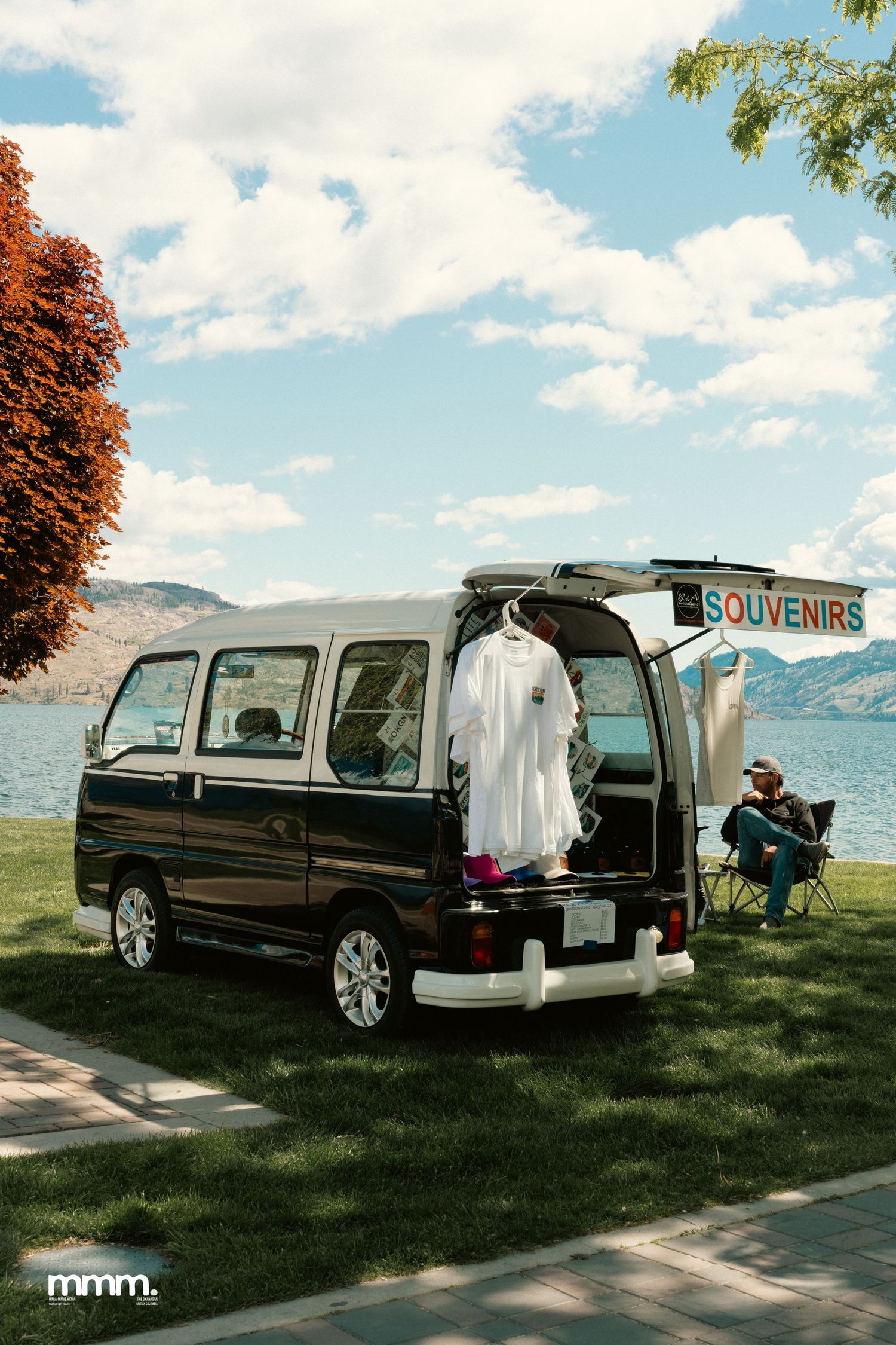Small black van with souvenirs sign open near lakeside on sunny day.