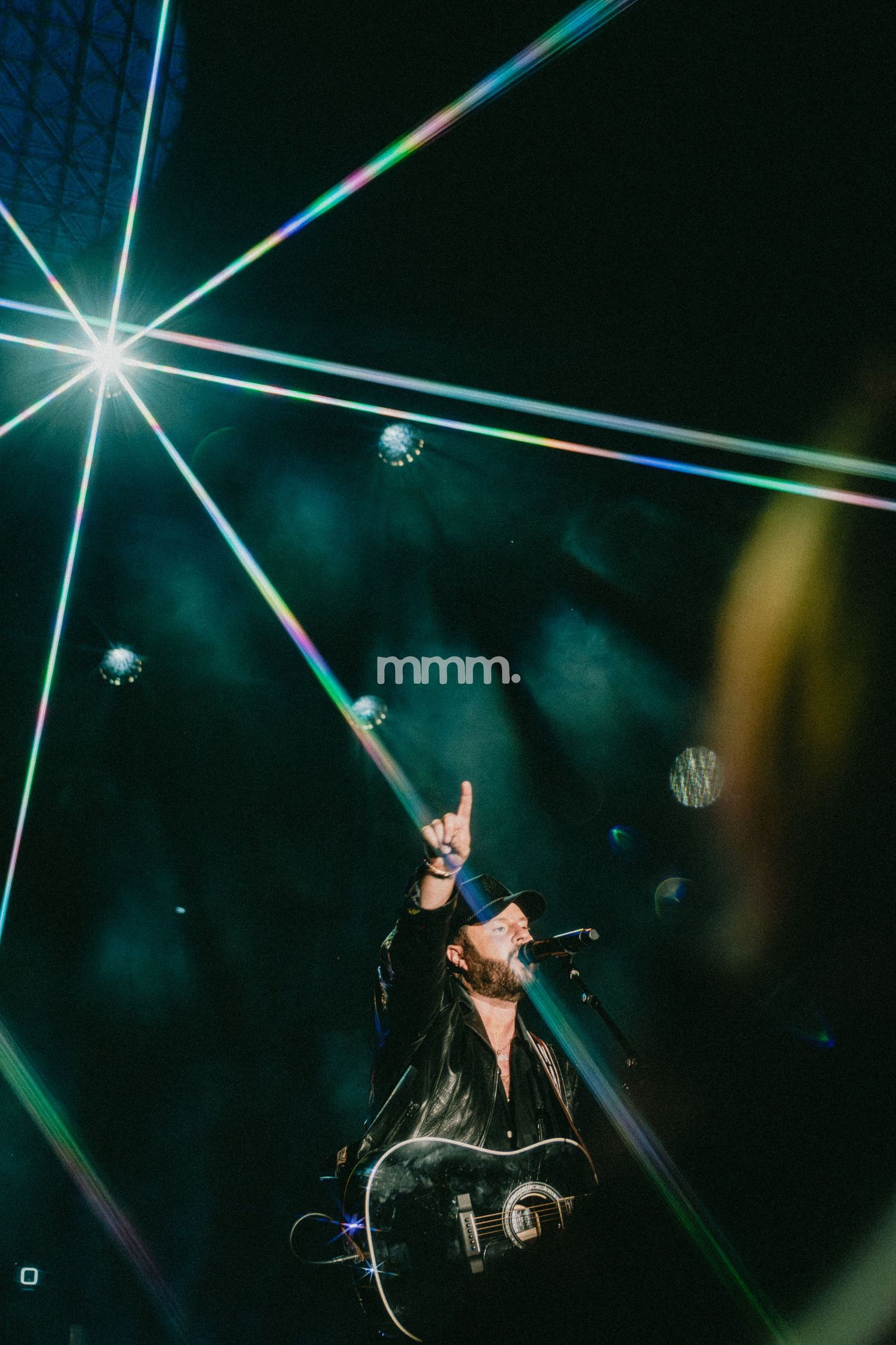 Guitarist performing on stage with dramatic rainbow laser lights and bokeh effects in a dark concert venue.