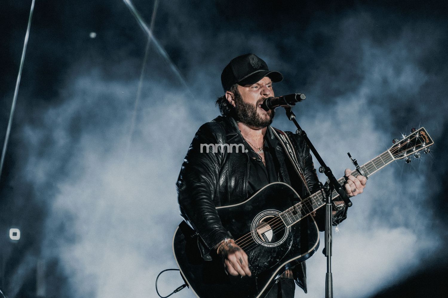 Male guitarist in black leather jacket and cap performs on smoky stage with acoustic guitar at live concert.