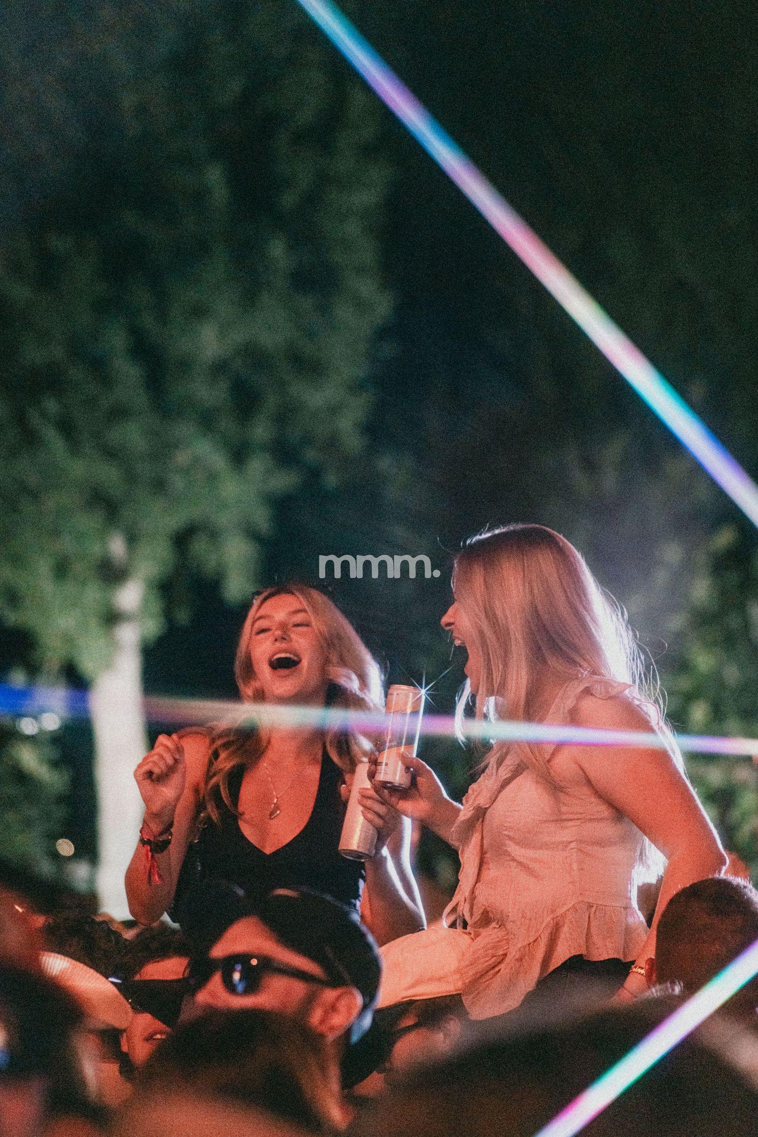 Two young women laugh and dance joyfully at an outdoor music festival at night, surrounded by colorful light beams.