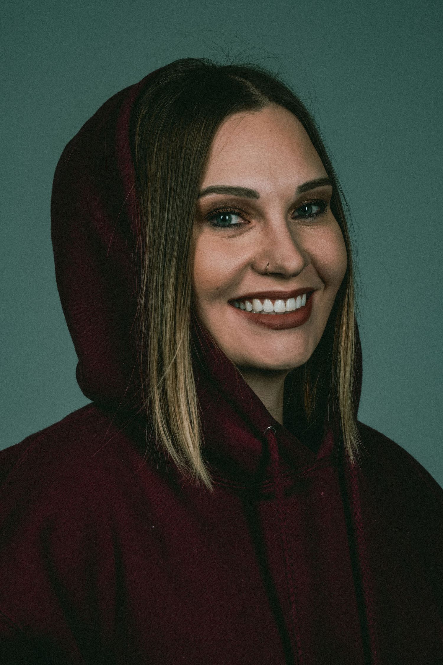 Happy woman with nose ring wearing a burgundy hoodie smiling against a teal background.