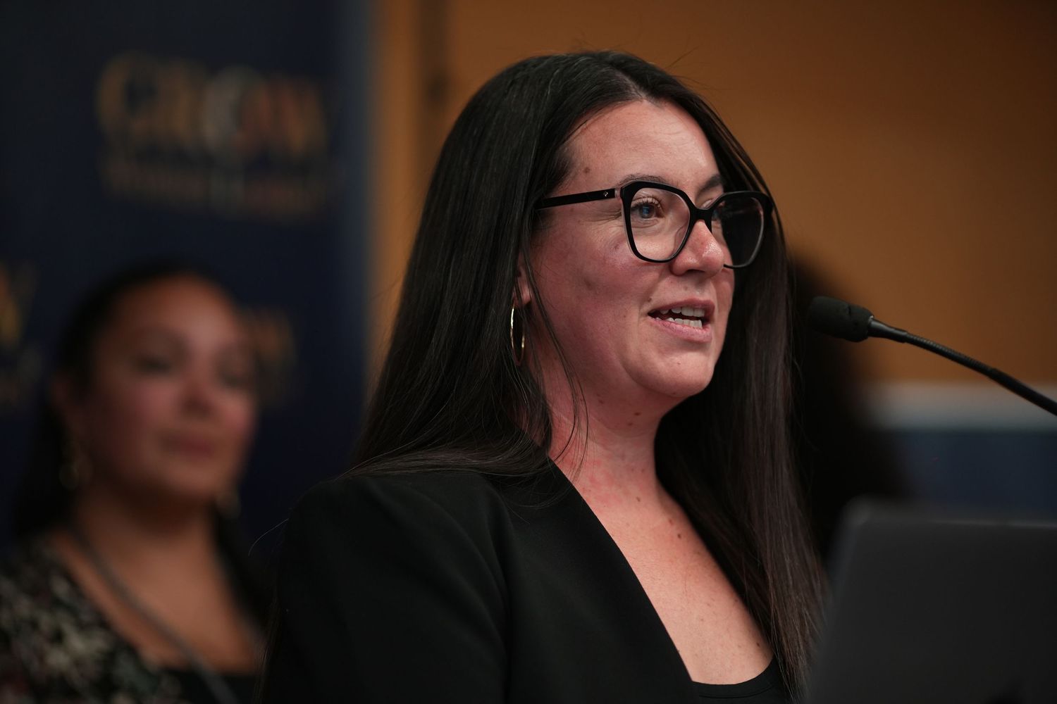 Woman with black hair and glasses speaking at a GROW Women Leaders event microphone.