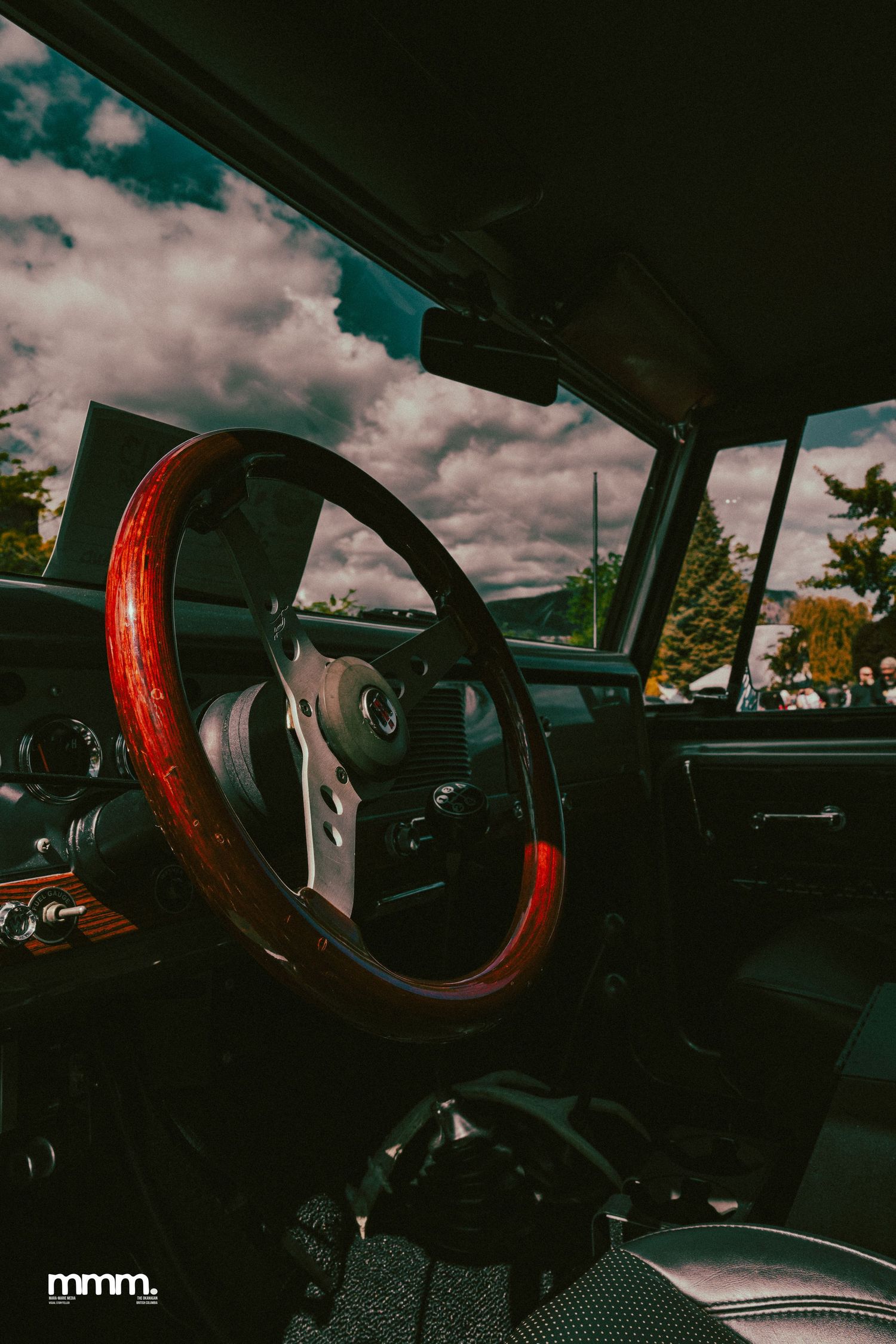 Vintage car interior with wood-grain steering wheel and cloudy sky view through windshield.