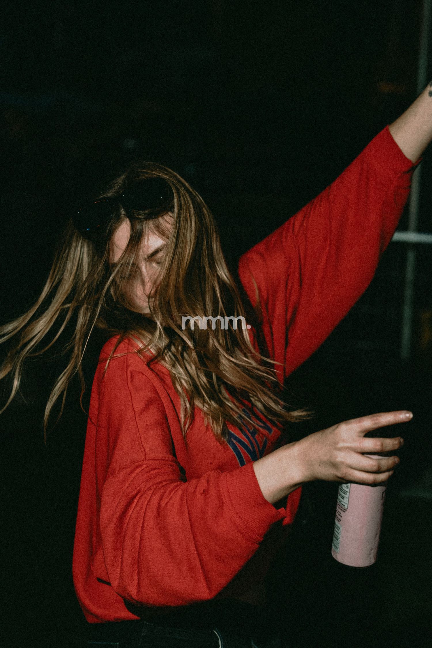 Young woman in red jacket dancing with arm raised, holding a pink cup, hair flowing, dark background.