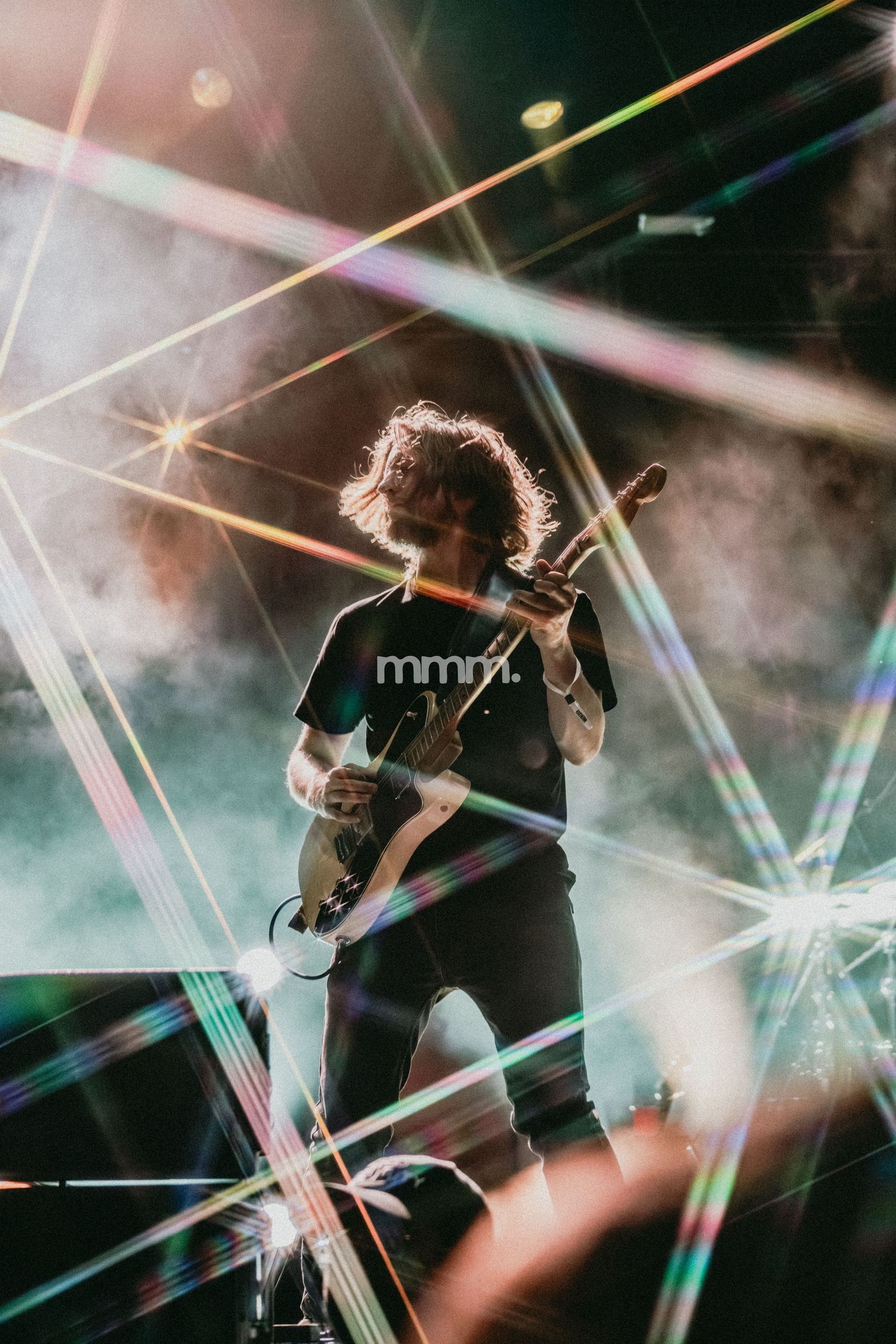 Guitarist performing on stage surrounded by colorful laser lights and smoke during a live concert.