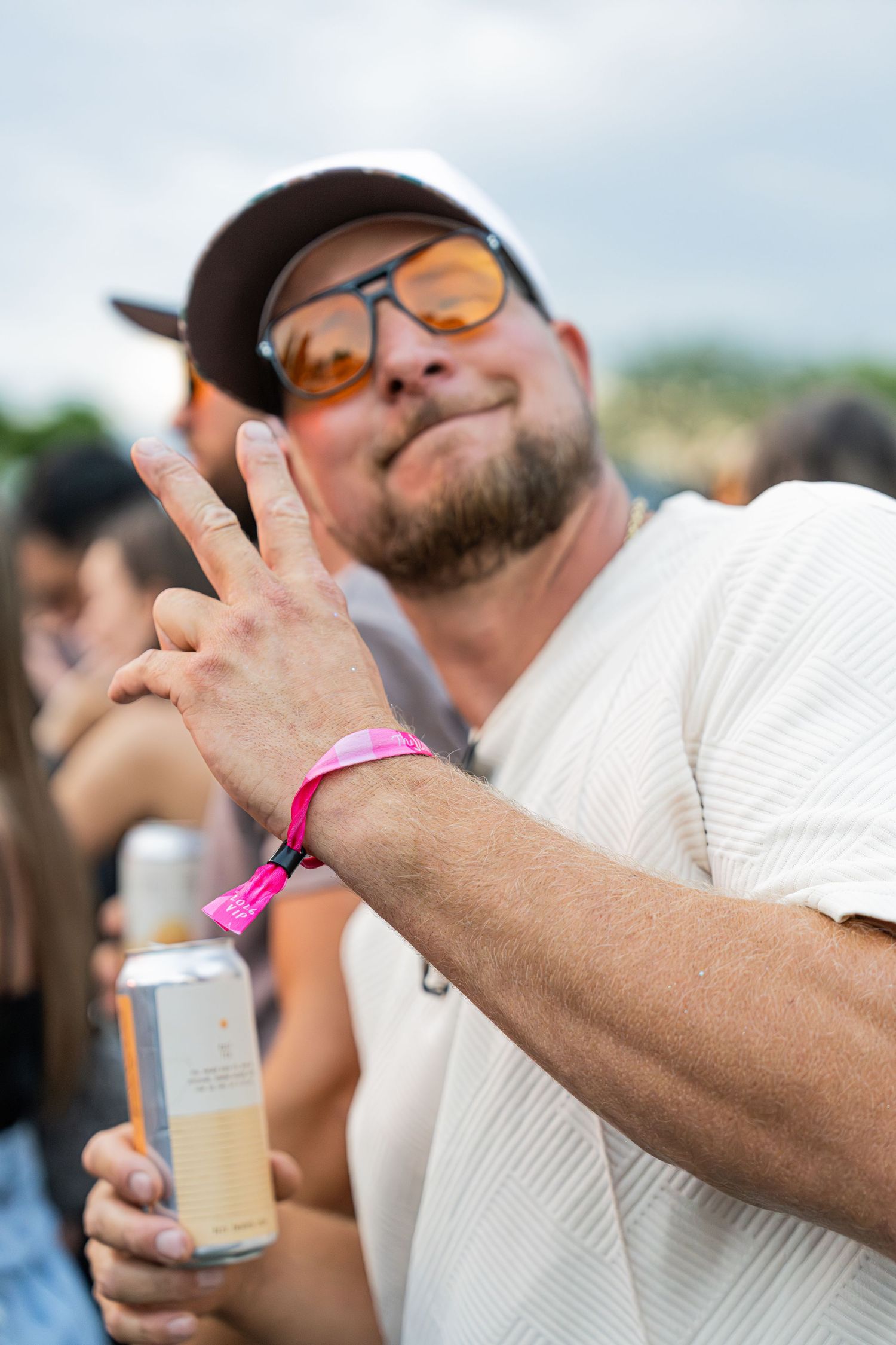 Bearded man in white shirt flashing peace sign while holding a drink at an outdoor festival.
