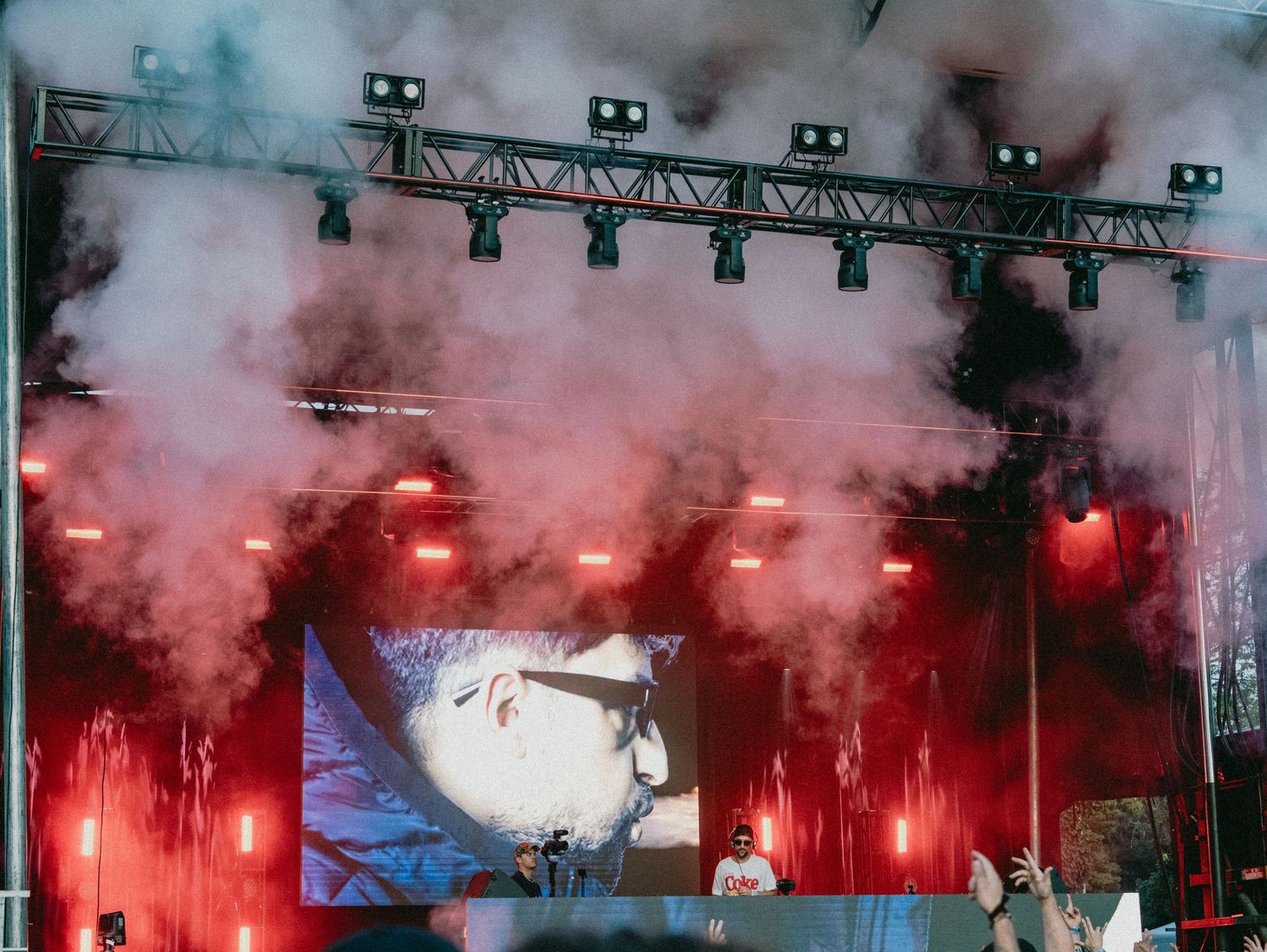A DJ performs on a smoke-filled festival stage with dramatic red lighting and pyrotechnics at an outdoor concert.