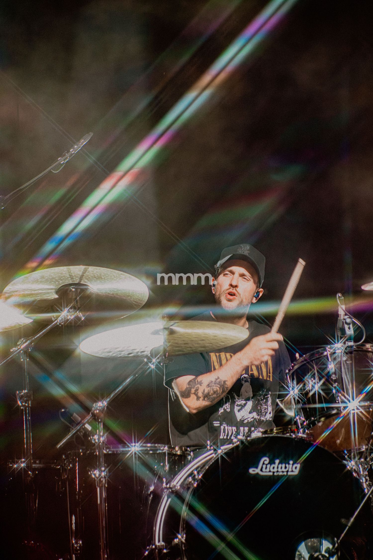 A tattooed drummer plays Ludwig drums on stage with colorful light refractions and sparkle effects during a live concert.