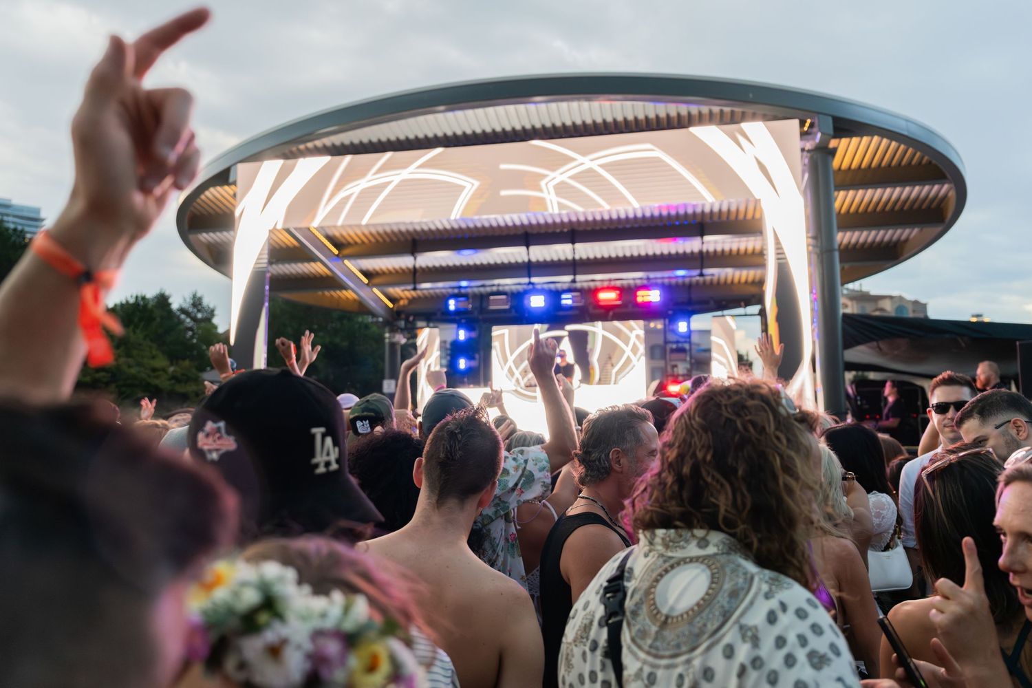 Festival crowd with raised hands facing an illuminated stage with colorful lights at an outdoor event.