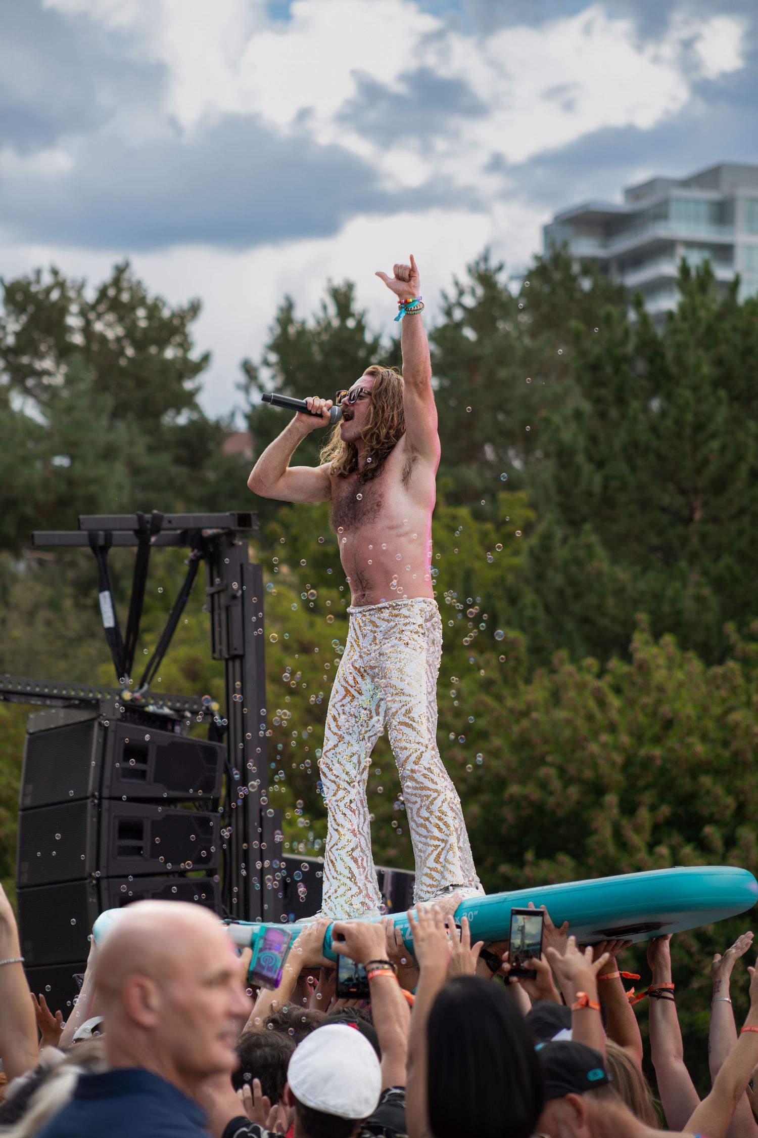 Shirtless performer in white pants crowd surfing on a surfboard at an outdoor music festival.