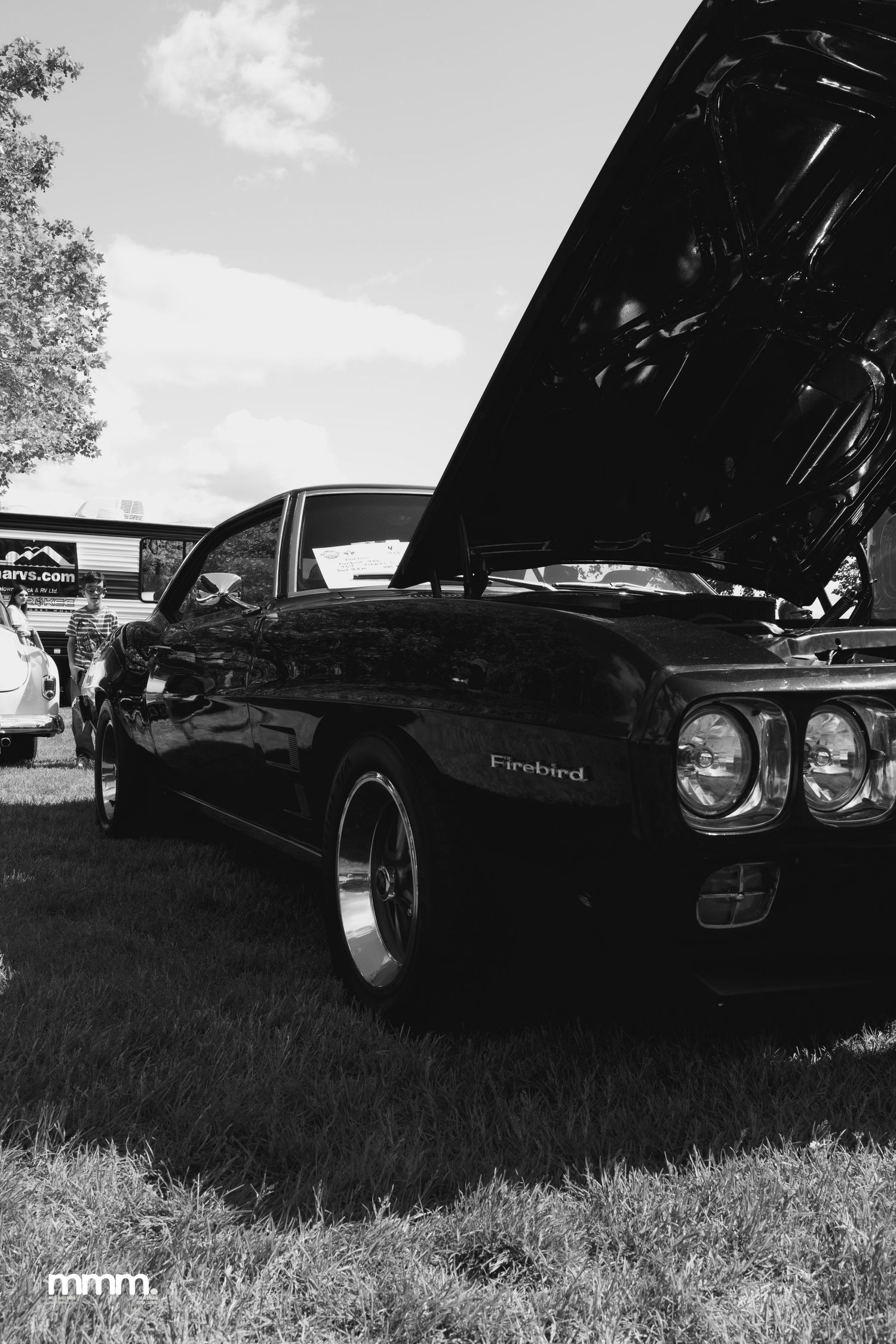 Classic Pontiac Firebird muscle car with hood open at outdoor car show, black and white photo.