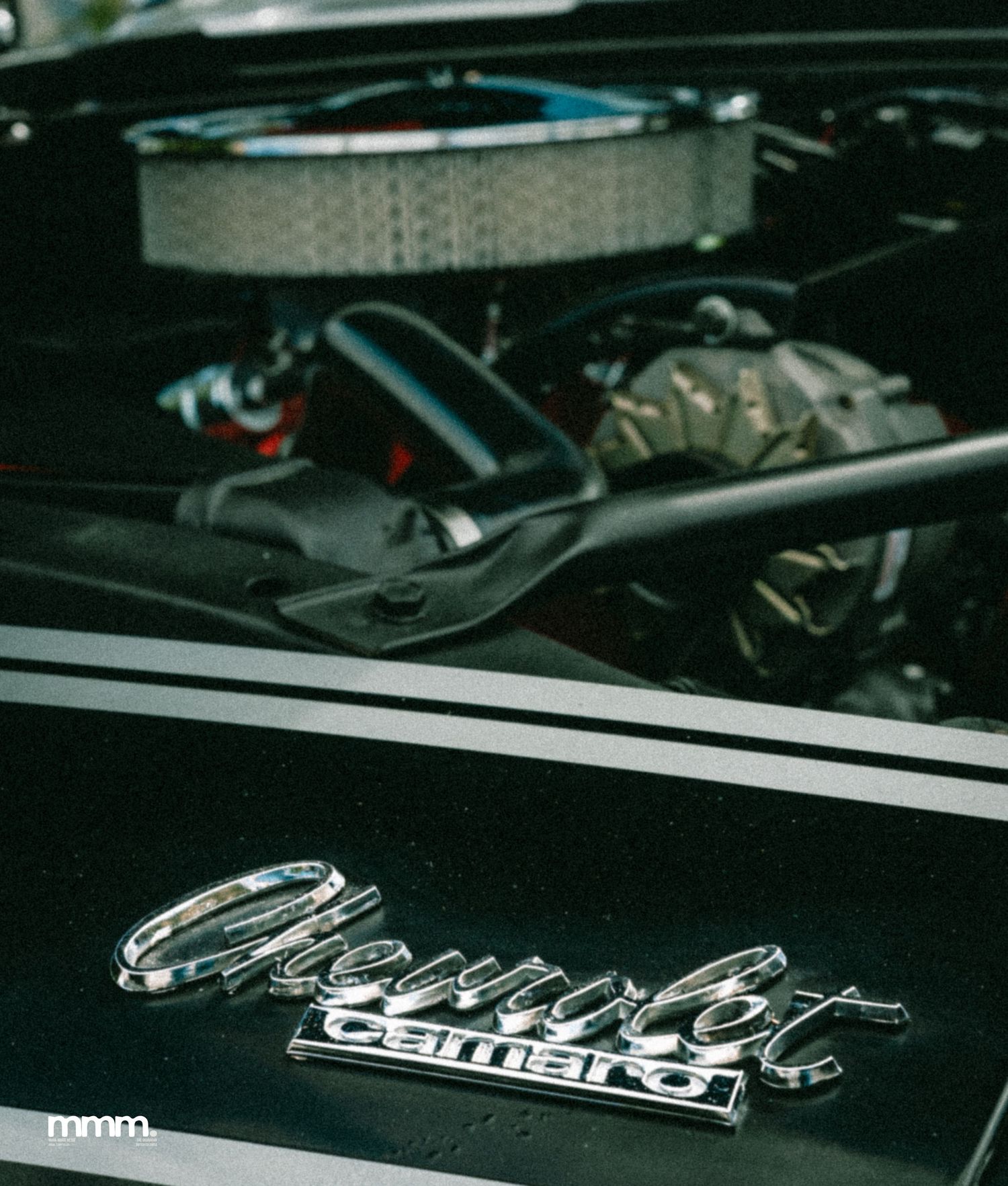 Chrome Chevrolet Camaro badge on black hood with engine visible.