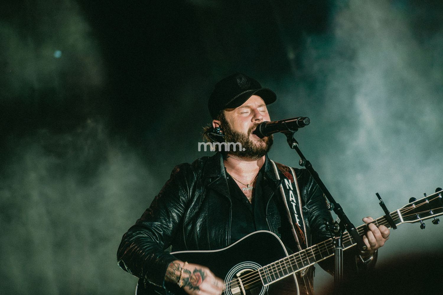 Tattooed male singer performing on stage with acoustic guitar, wearing black jacket and cap, with dramatic fog lighting.