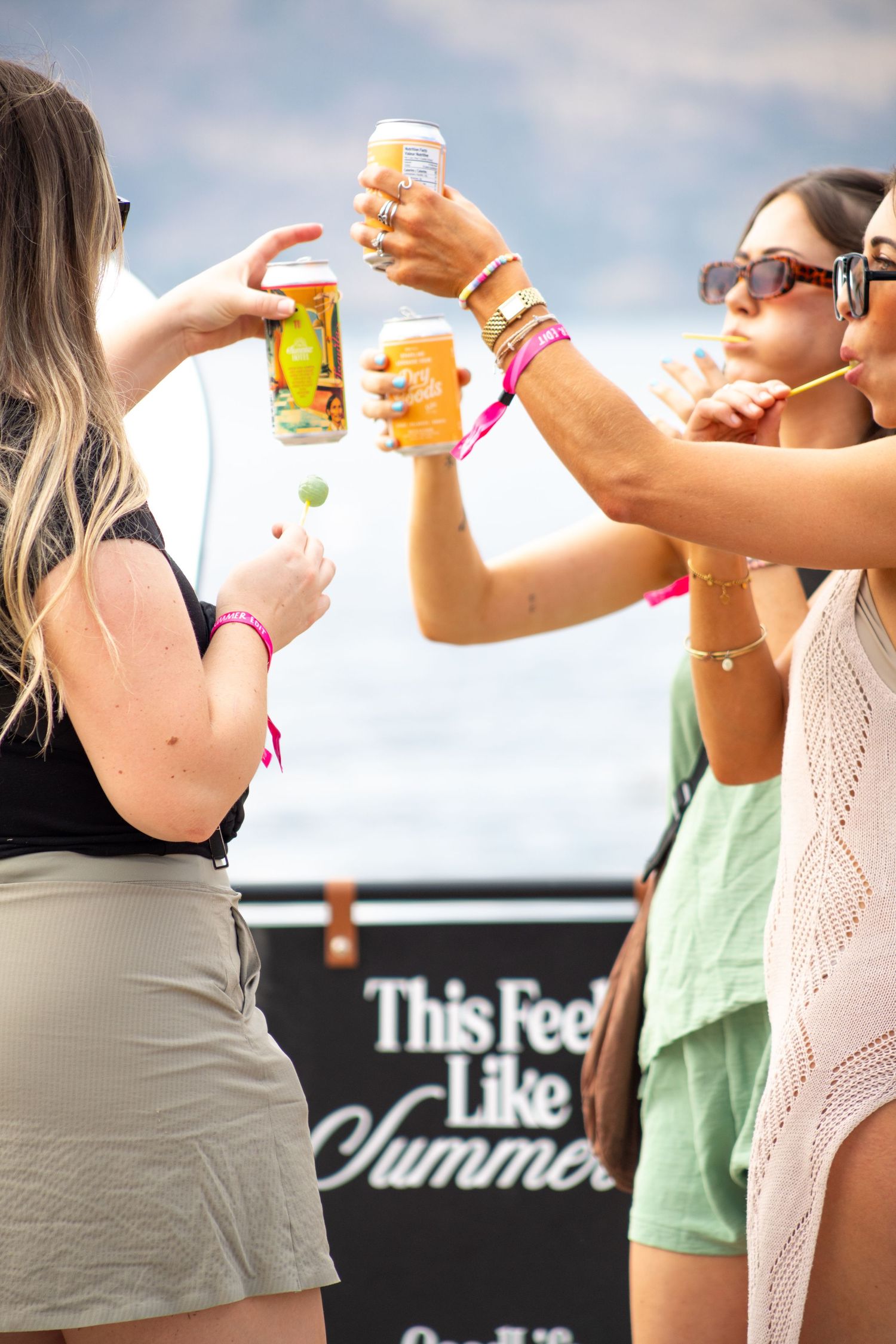 Group of women toasting drinks at a summer music festival near a This Feels Like Summer banner.